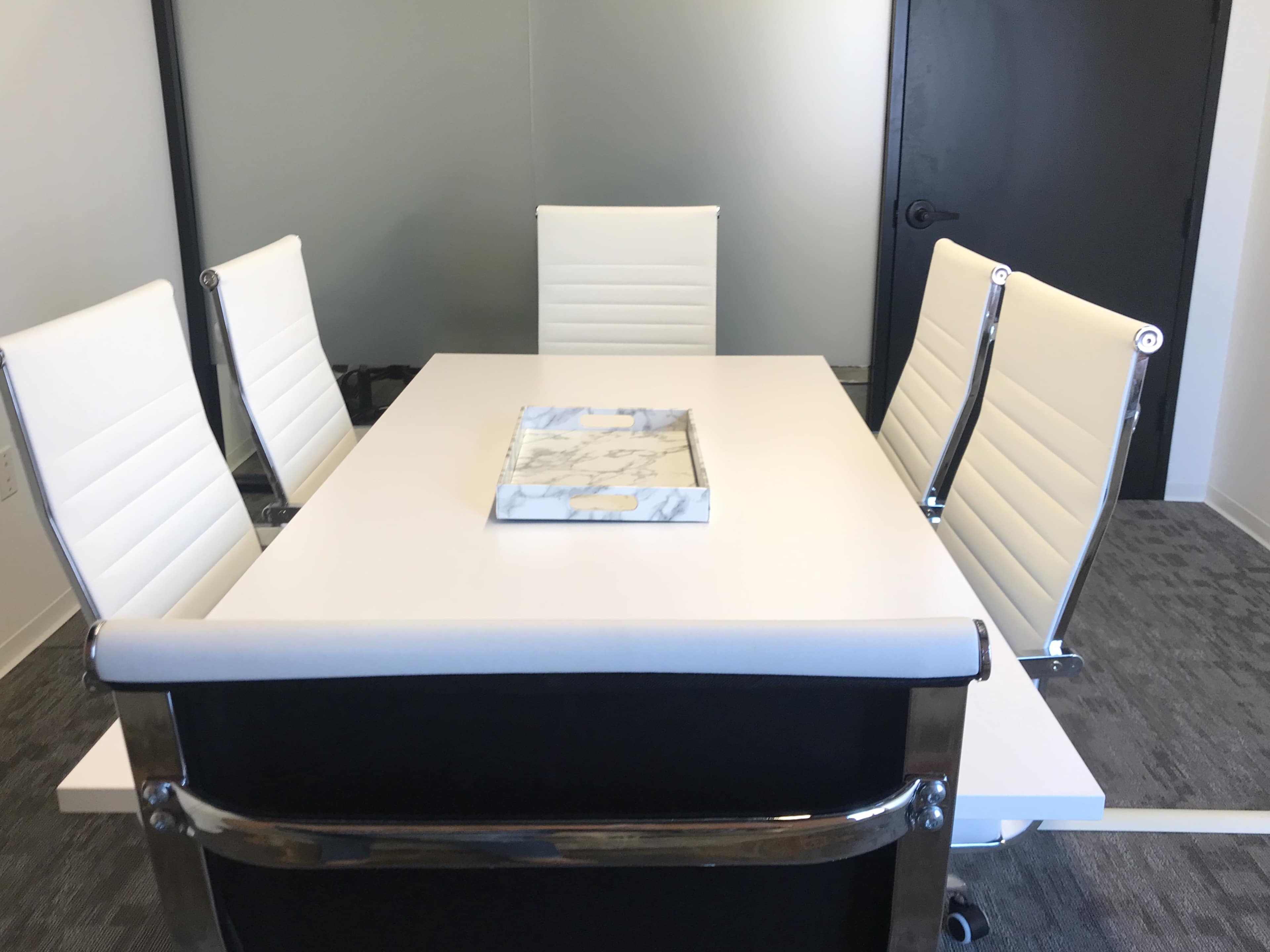 A modern conference room features a white table surrounded by four white leather chairs and a marble-patterned tray in the center.