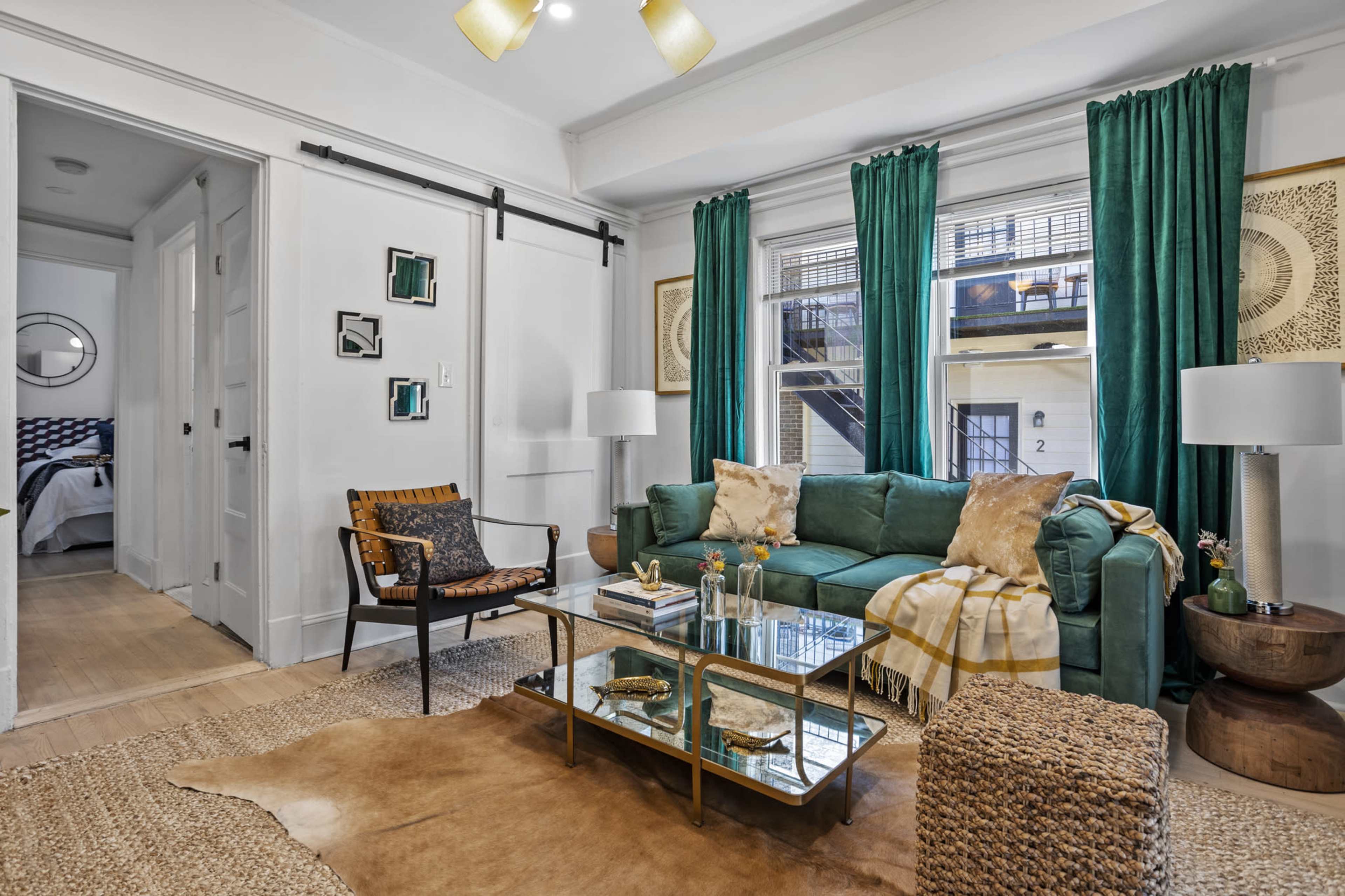 The living room features a green sofa, glass coffee table, and patterned curtains, with neutral décor elements and a view into another room through an open doorway.
