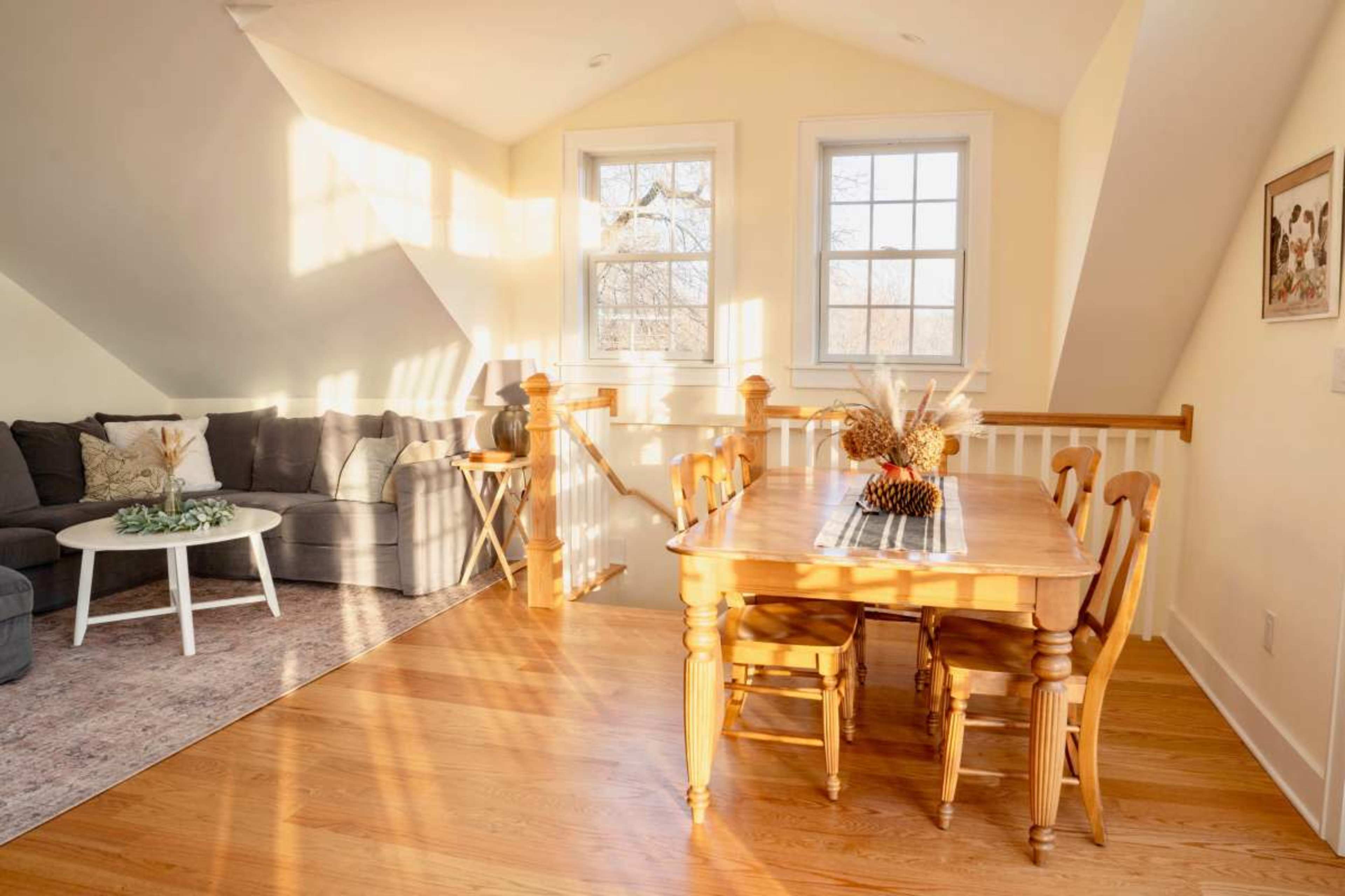 A bright, cozy living space features a gray sectional sofa, a small round table, and a wooden dining table with chairs, illuminated by sunlight filtering through the windows.