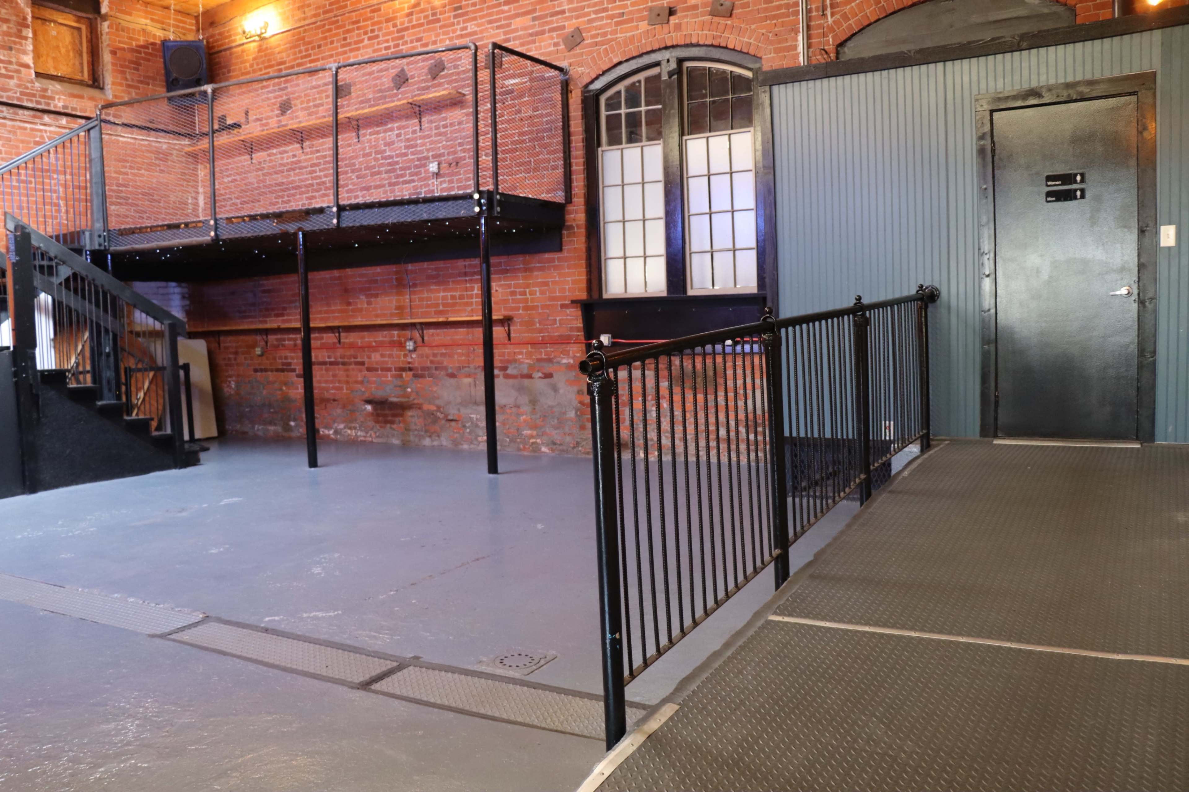 The image shows an industrial-style interior space with exposed brick walls, a staircase leading to a raised platform, and a metal railing along a flat surface.