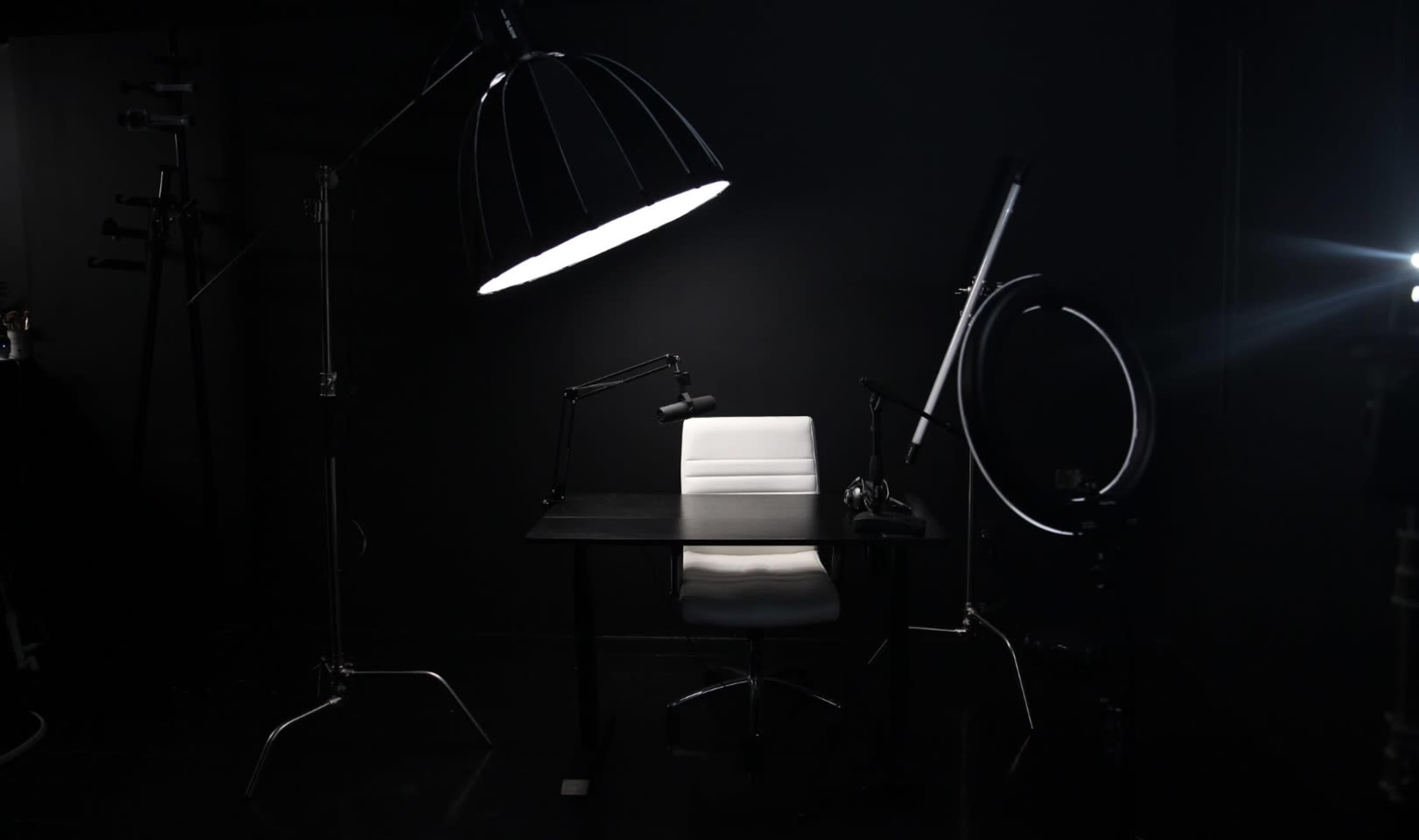 A well-lit photography studio setup features a white chair next to a black desk under large light fixtures against a dark backdrop.