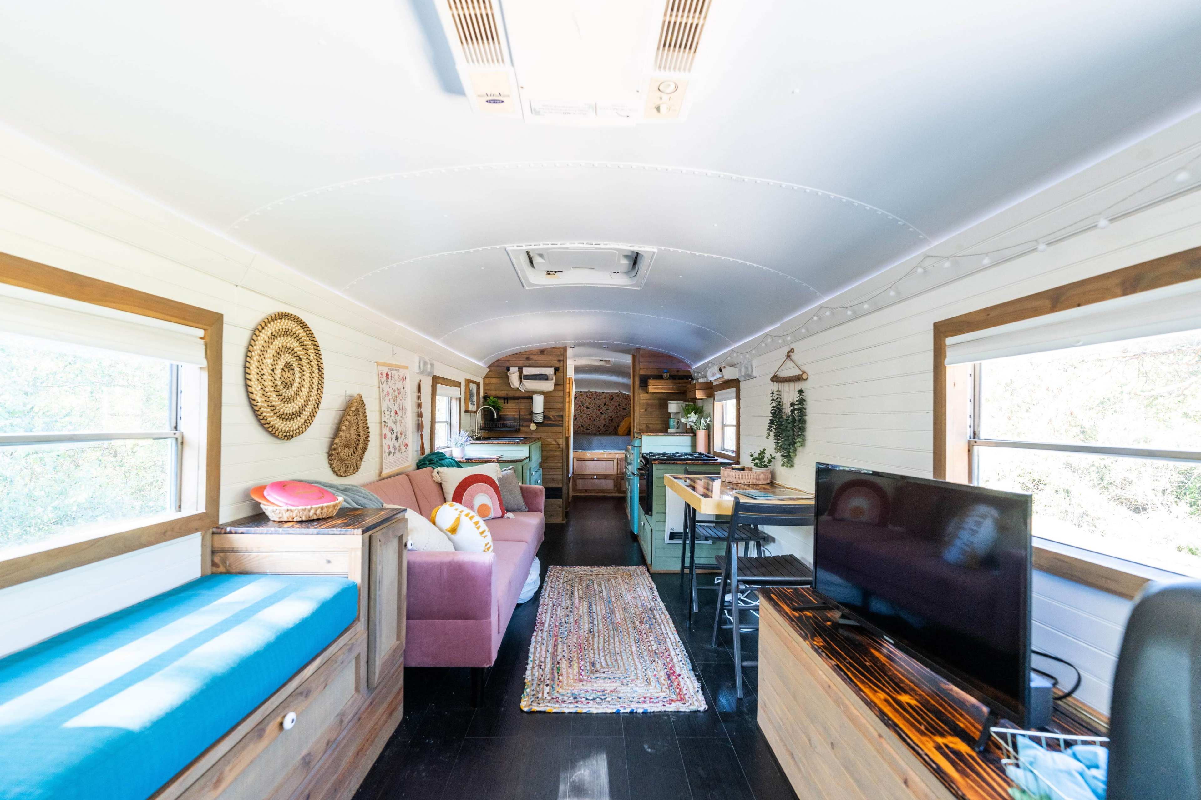 The image shows the interior of a renovated bus, featuring a living area with a blue couch, a pink sofa, a kitchen area, and large windows that provide natural light.