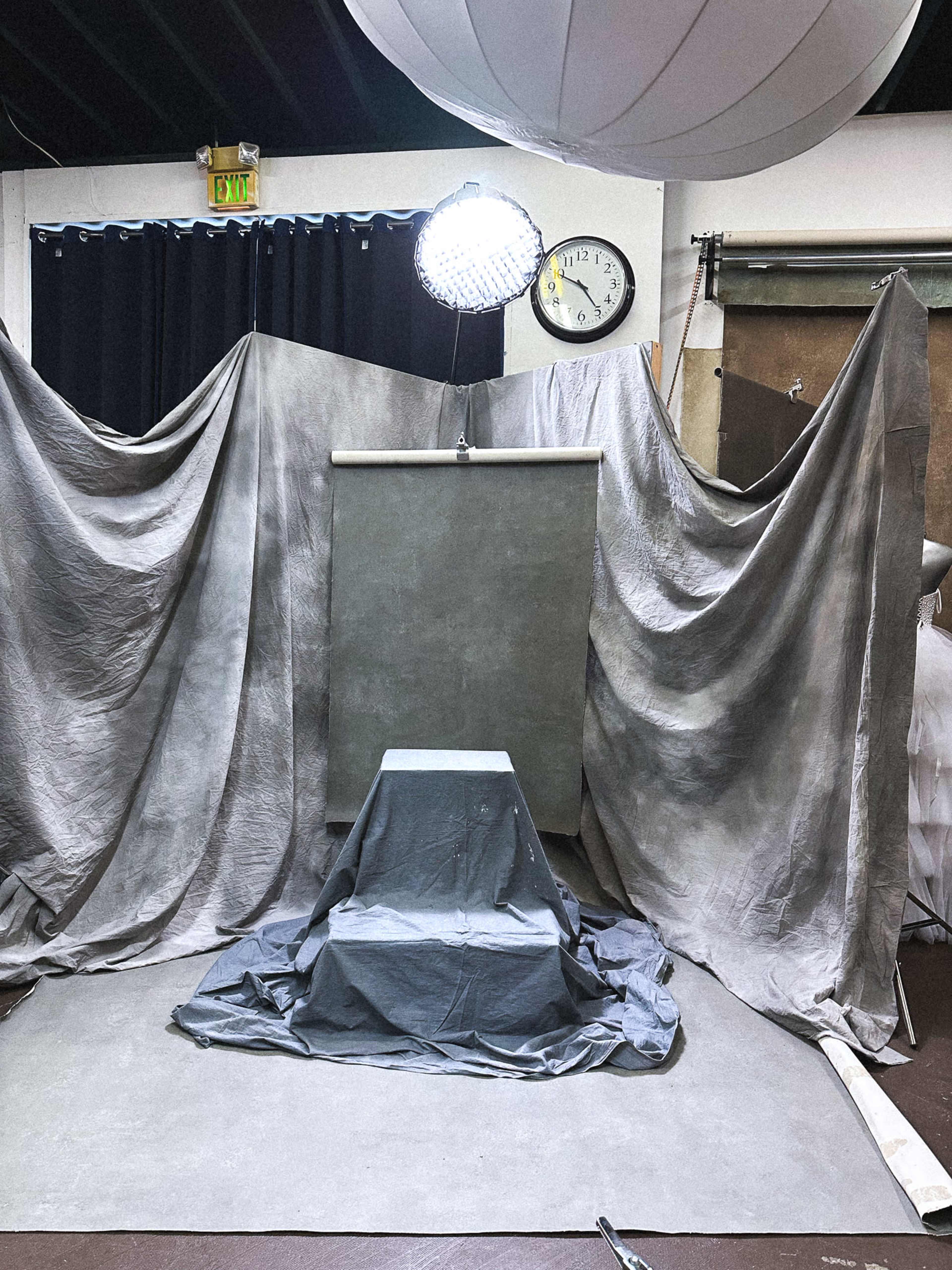 The image shows a photography studio setup with draped gray fabrics, a backdrop, and a spotlight illuminating a pedestal covered with a gray cloth.