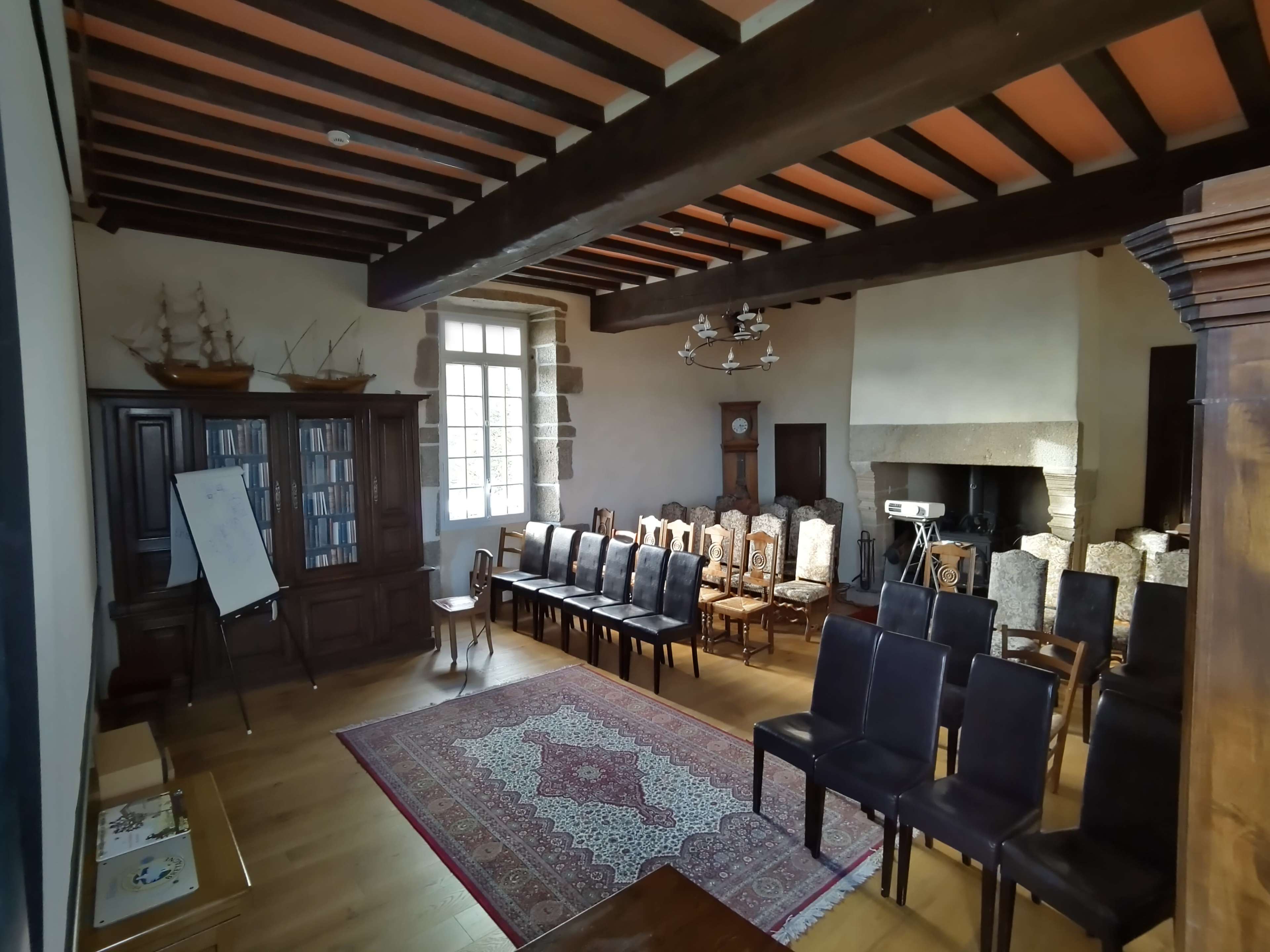 The image shows a large, organized room with wooden beams, rows of chairs, and a fireplace, featuring a bookshelf and a whiteboard.