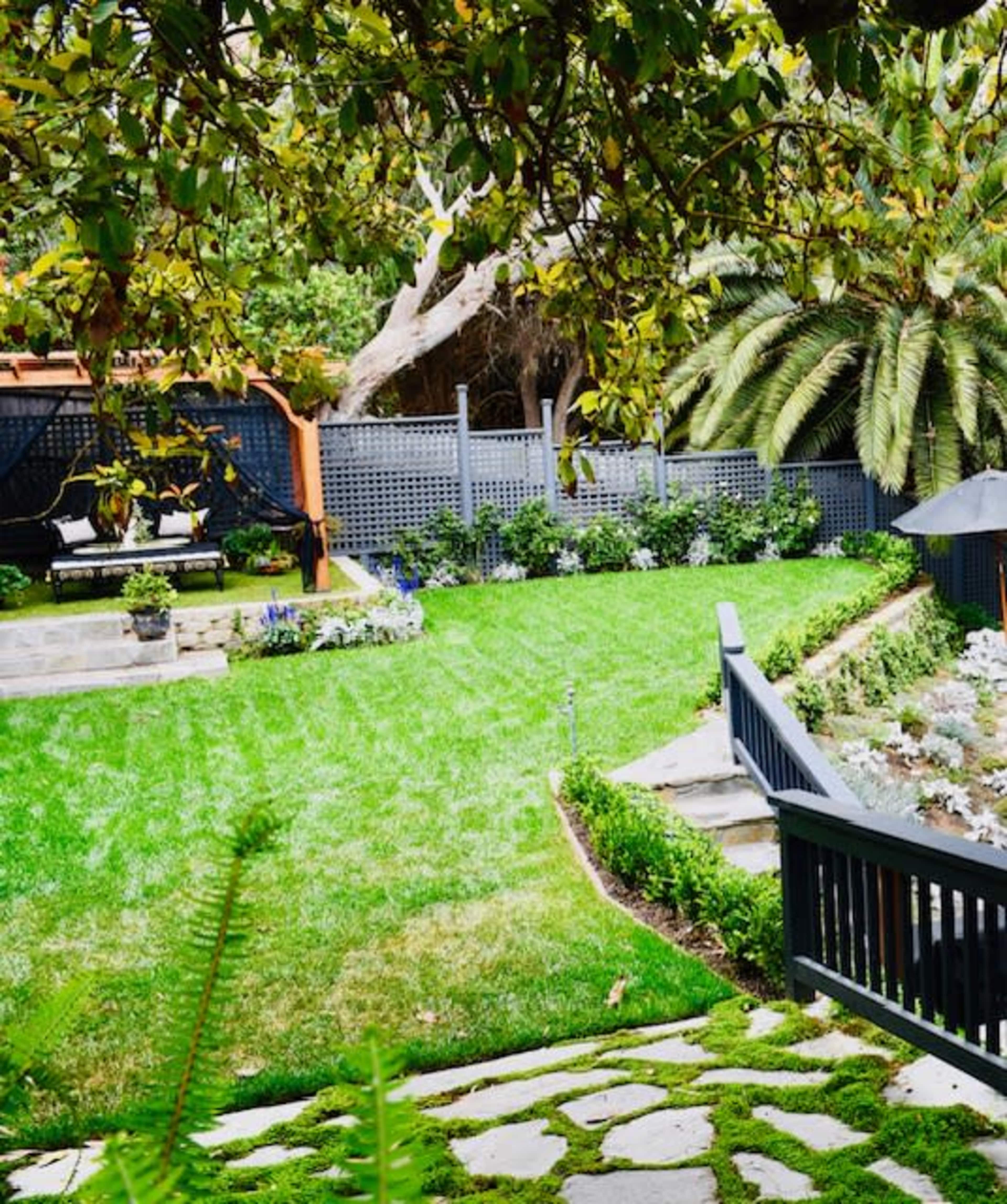 A well-maintained backyard features lush green grass, stone pathways, and a seating area under a pergola surrounded by various plants.
