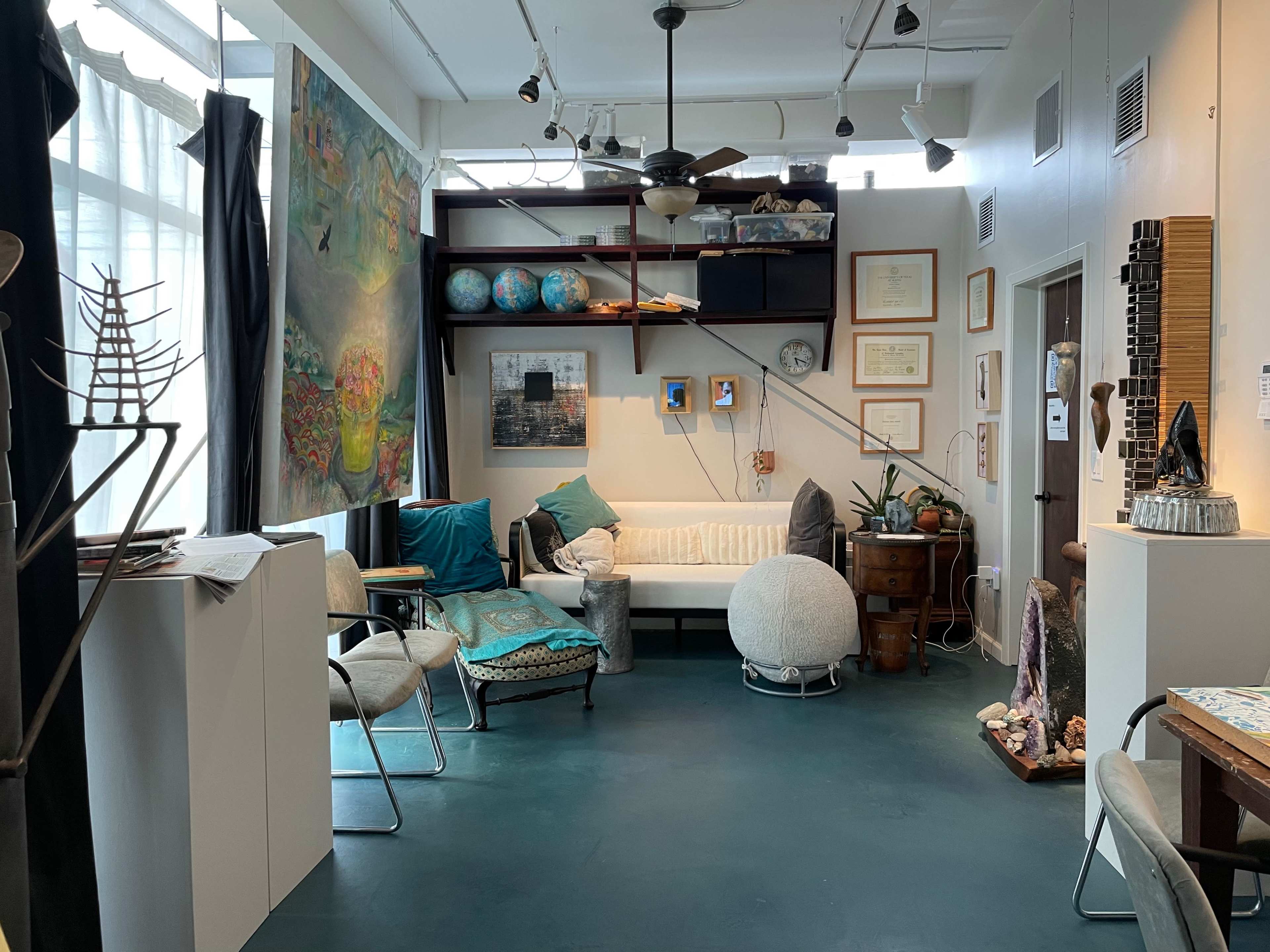 The image shows a cozy studio or office space featuring various seating options, artwork on the walls, and decorative globes on a bookshelf.