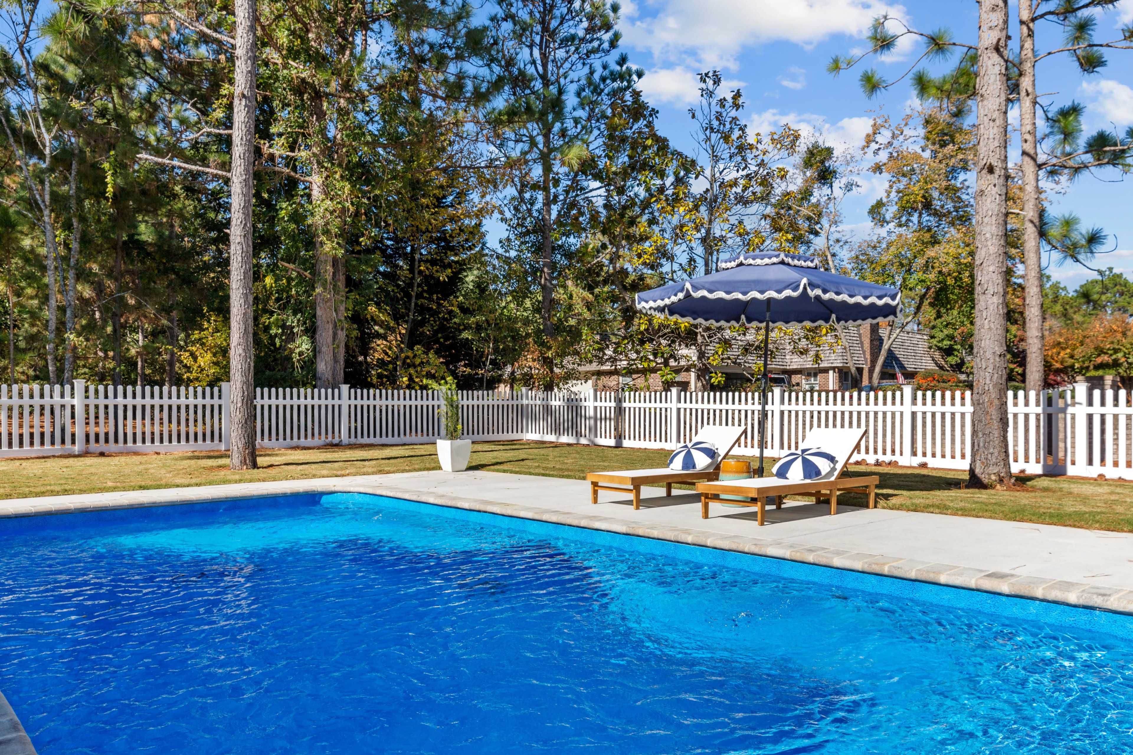 A blue swimming pool with lounge chairs and an umbrella is surrounded by trees and a white picket fence.