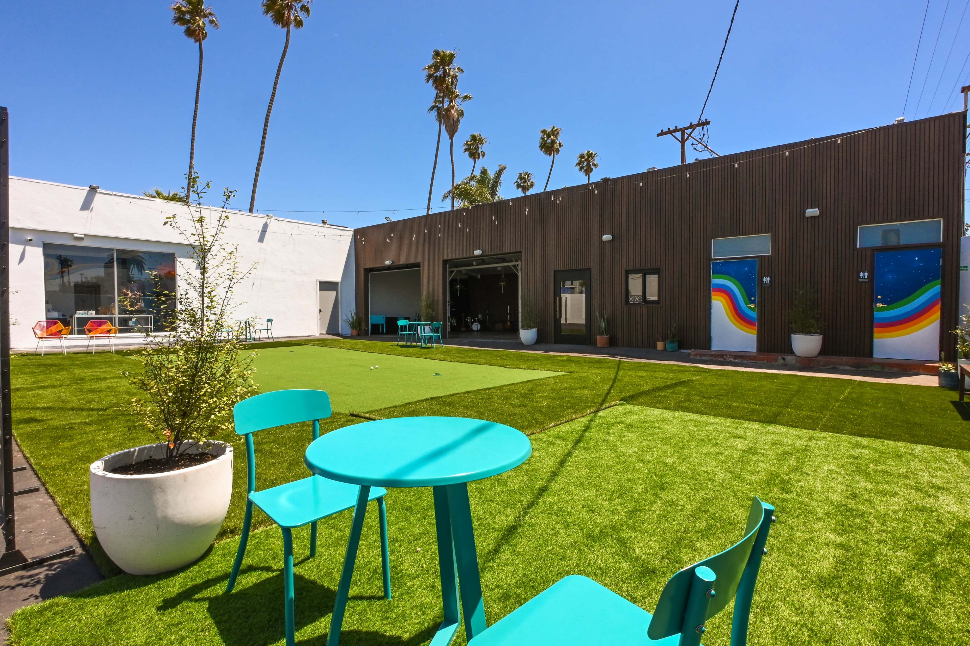 A spacious outdoor area featuring artificial grass, two turquoise chairs, and buildings with colorful murals and palm trees in the background.