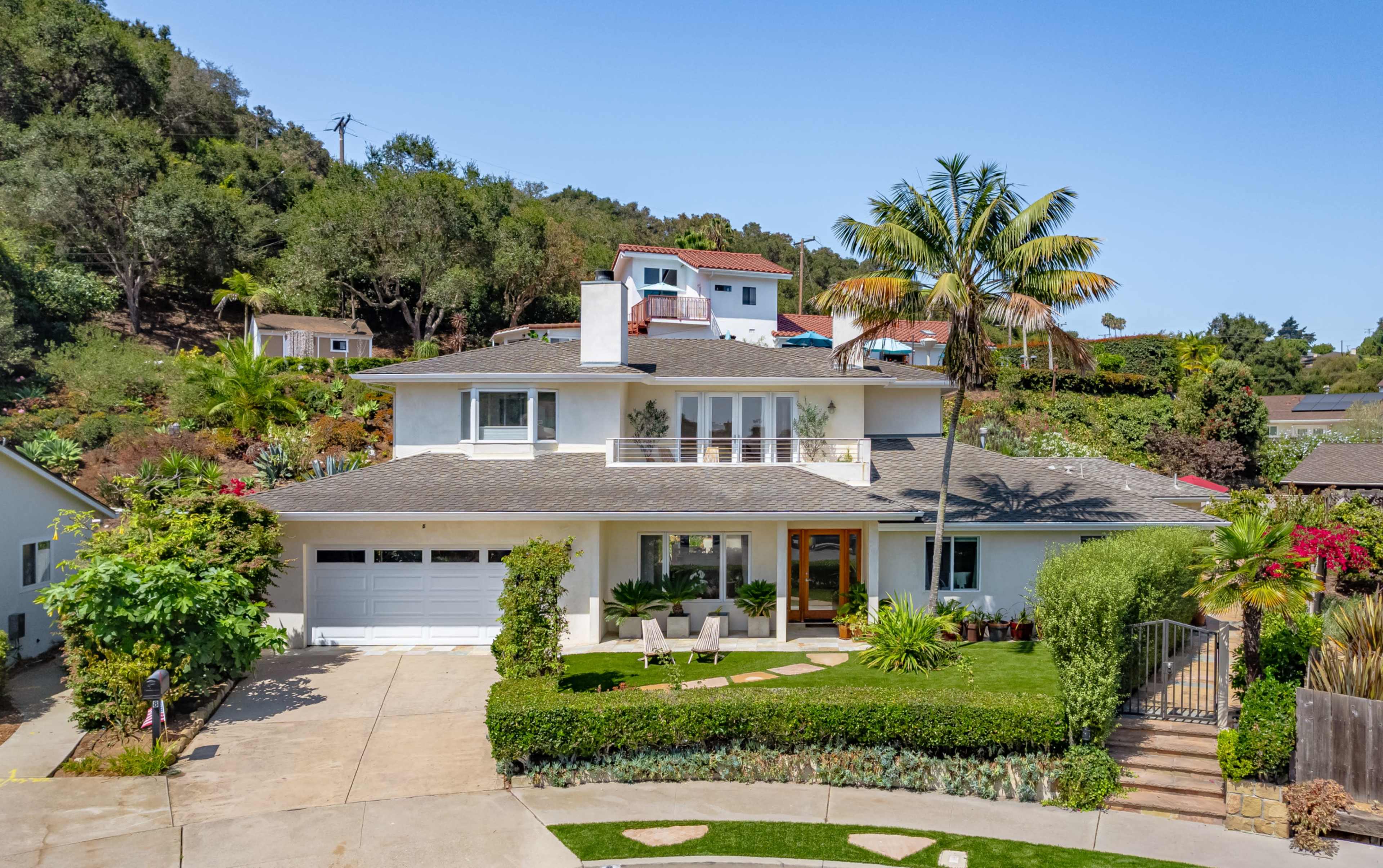 The image shows a large, two-story house with a landscaped front yard, a driveway, and palm trees surrounding the property.