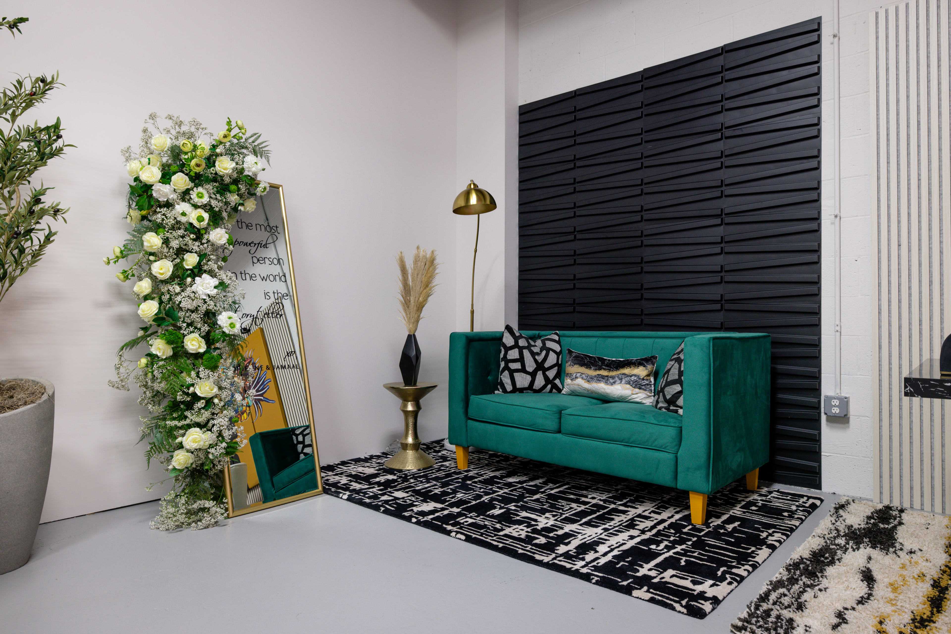 A green sofa with patterned cushions, a decorative mirror surrounded by flowers, a gold floor lamp, a potted plant, and a black-and-white patterned rug in a modern indoor setting.