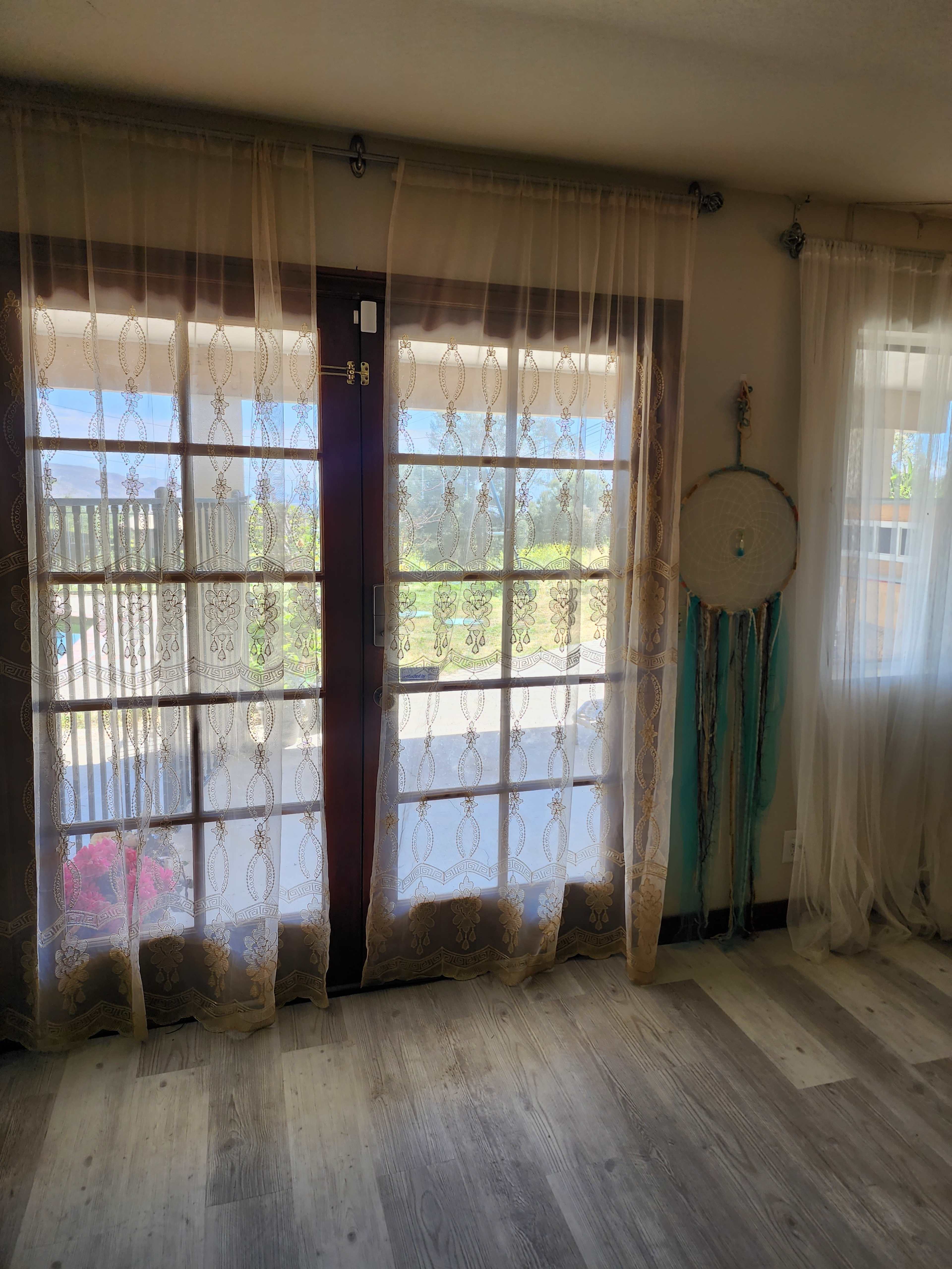 The image shows a room with sheer, patterned curtains over double glass doors leading to an outdoor view, and a dreamcatcher hanging nearby.