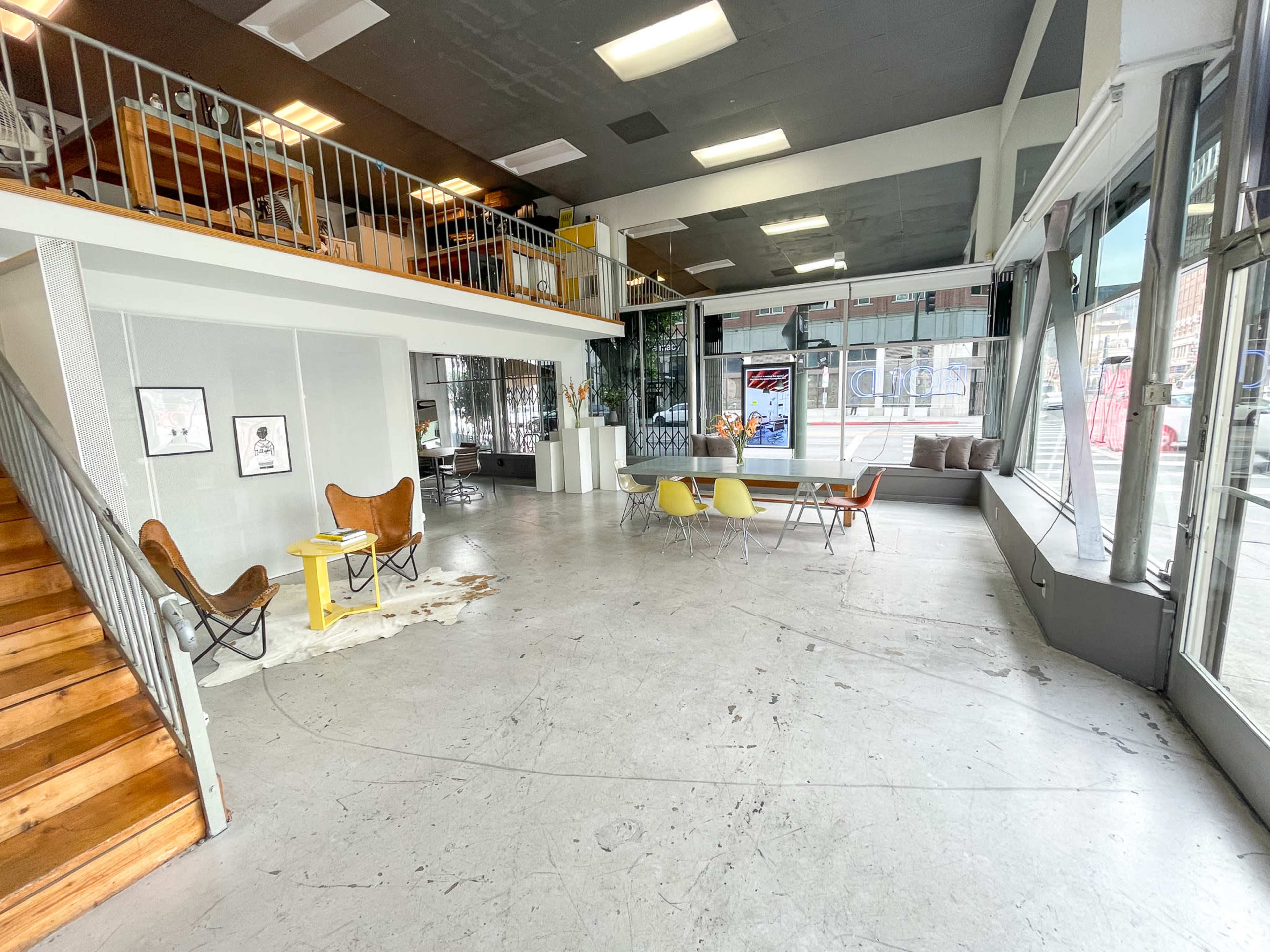 The image shows a spacious, modern interior of a storefront or gallery with a staircase, furniture arranged for seating, and large windows allowing natural light to enter.