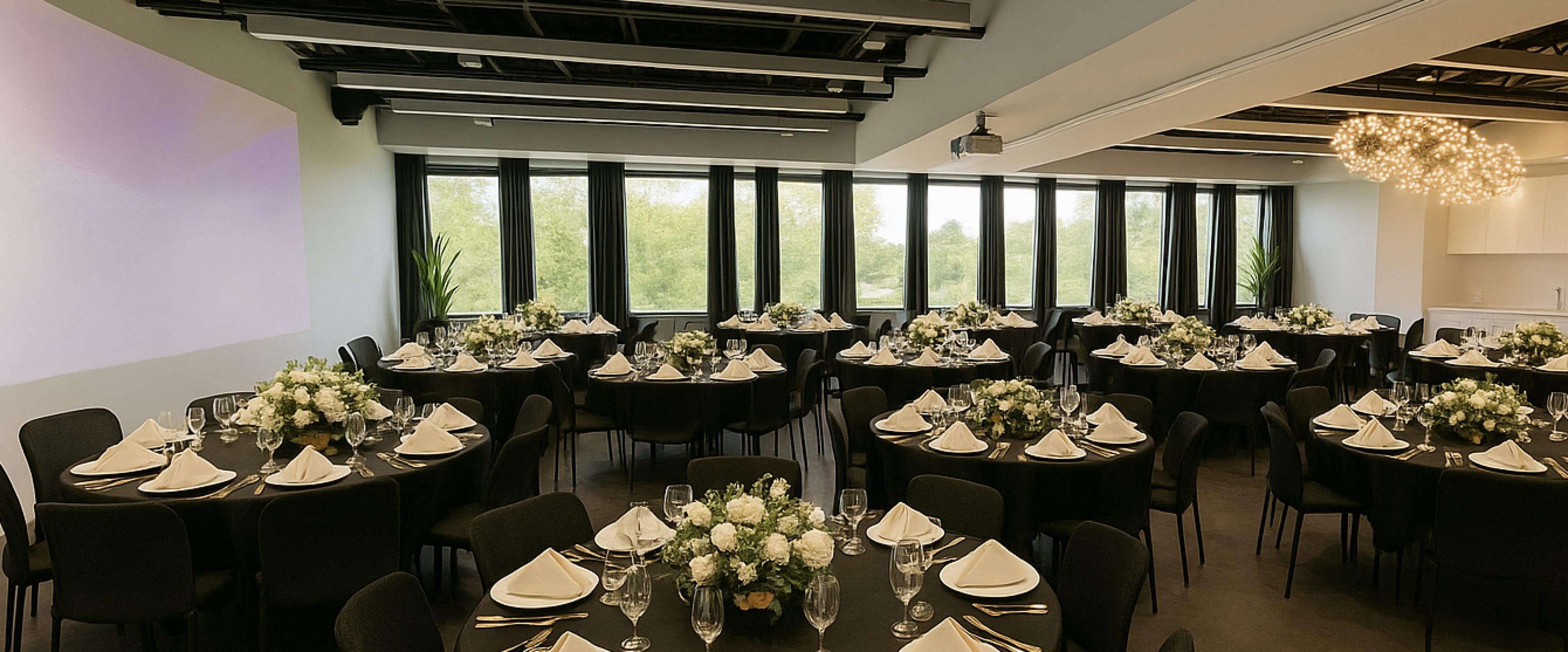 A banquet hall features round tables adorned with floral centerpieces, neatly arranged with white napkins and glassware, alongside large windows offering a view of greenery.