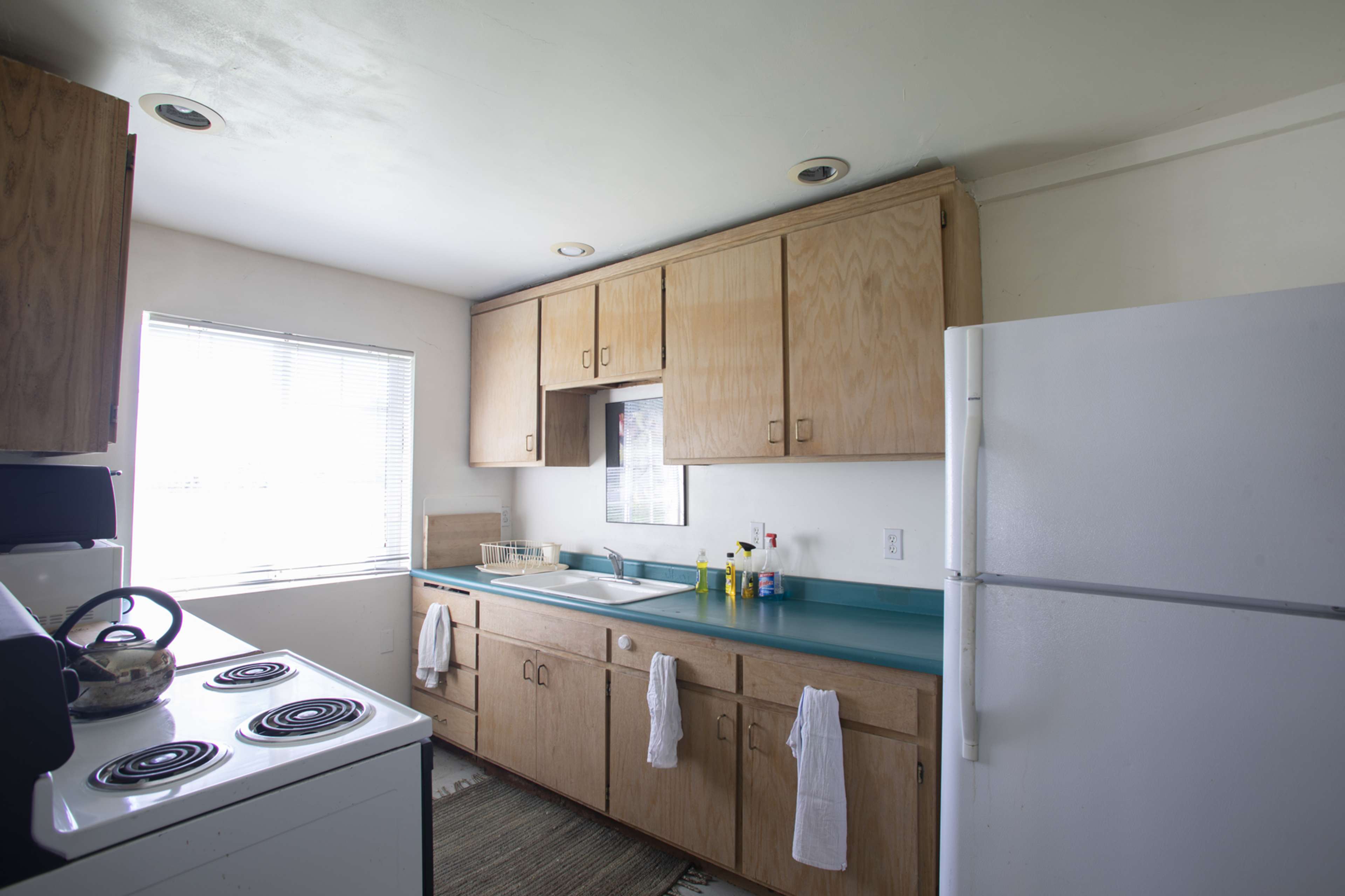 A simple kitchen with wooden cabinets, a white refrigerator, a sink, and a stove, along with several cleaning supplies on the countertop.