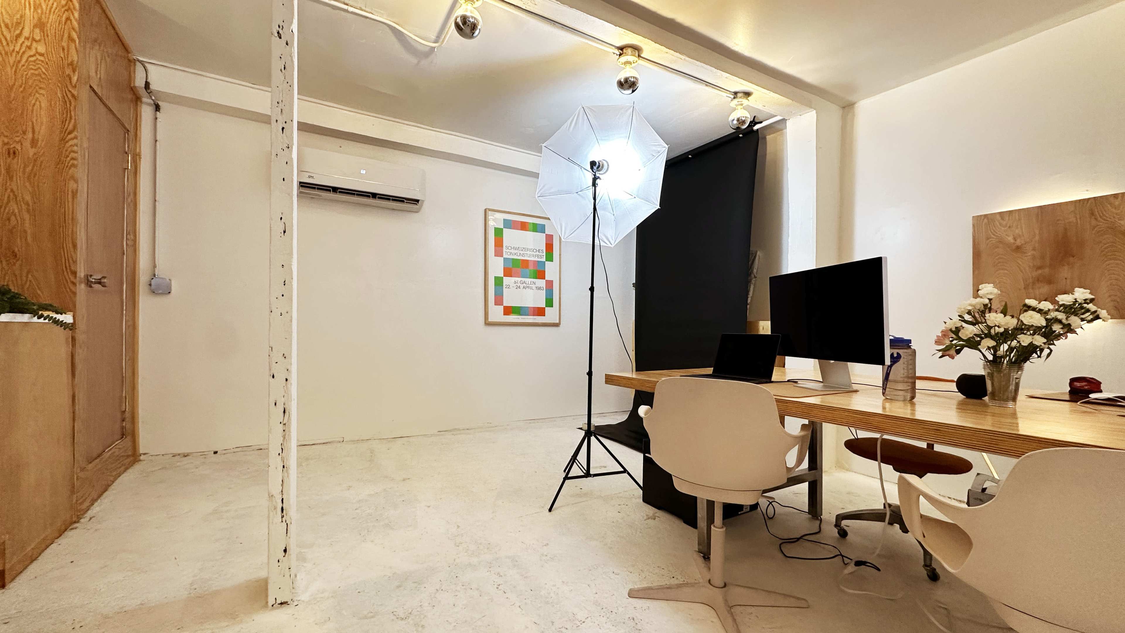A minimalist office space with a wooden table, chairs, a computer setup, and a photography lighting umbrella in a corner.