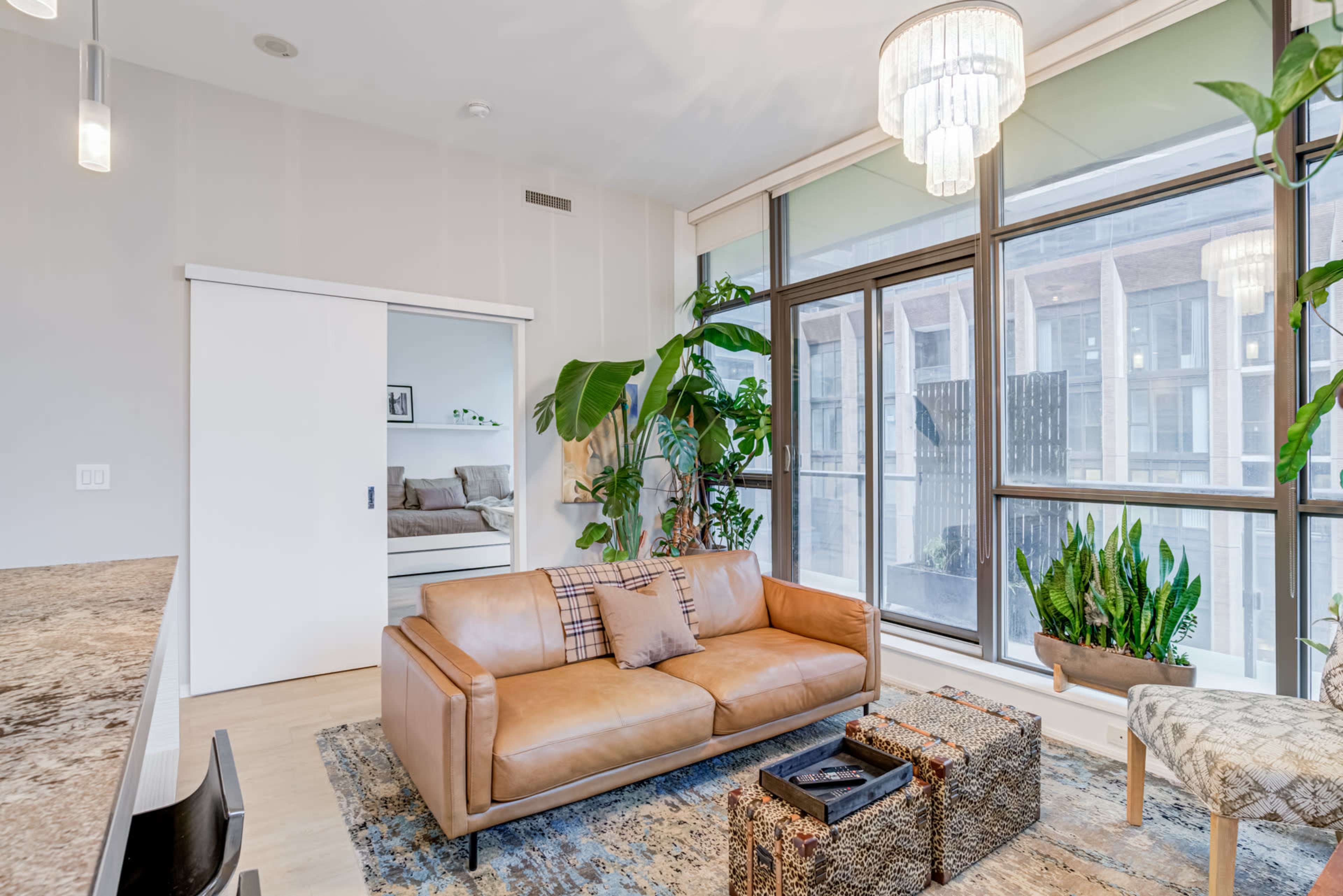 A modern living space features a leather sofa, large windows with greenery, and a light-colored rug.