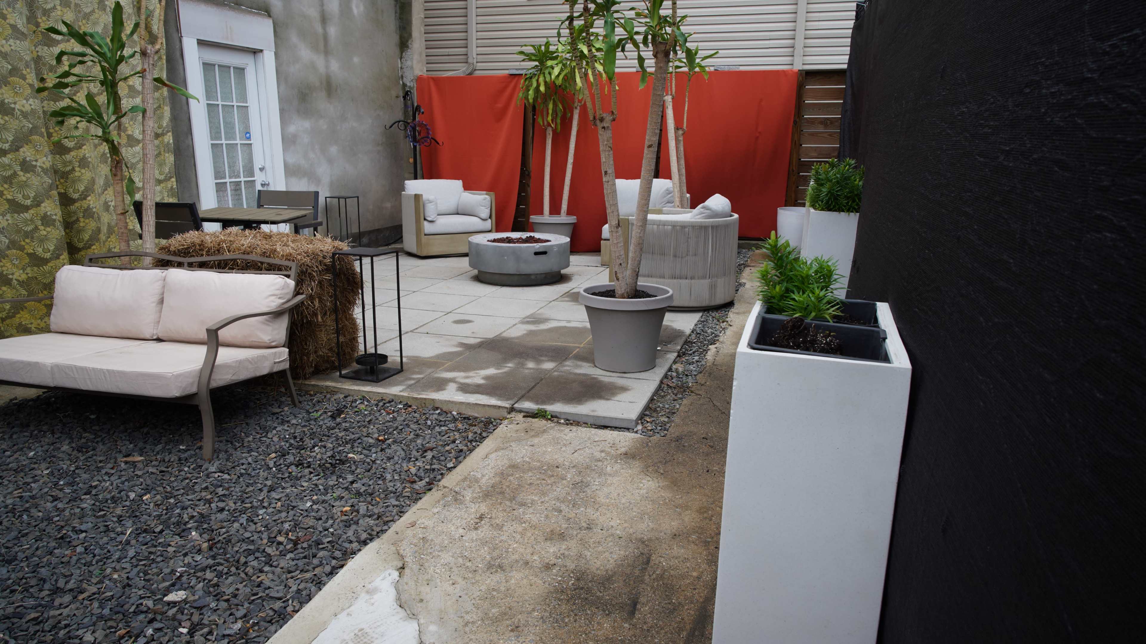 The image shows a small outdoor patio area featuring seating arrangements, planters, and a central fire pit, surrounded by walls and a red backdrop.