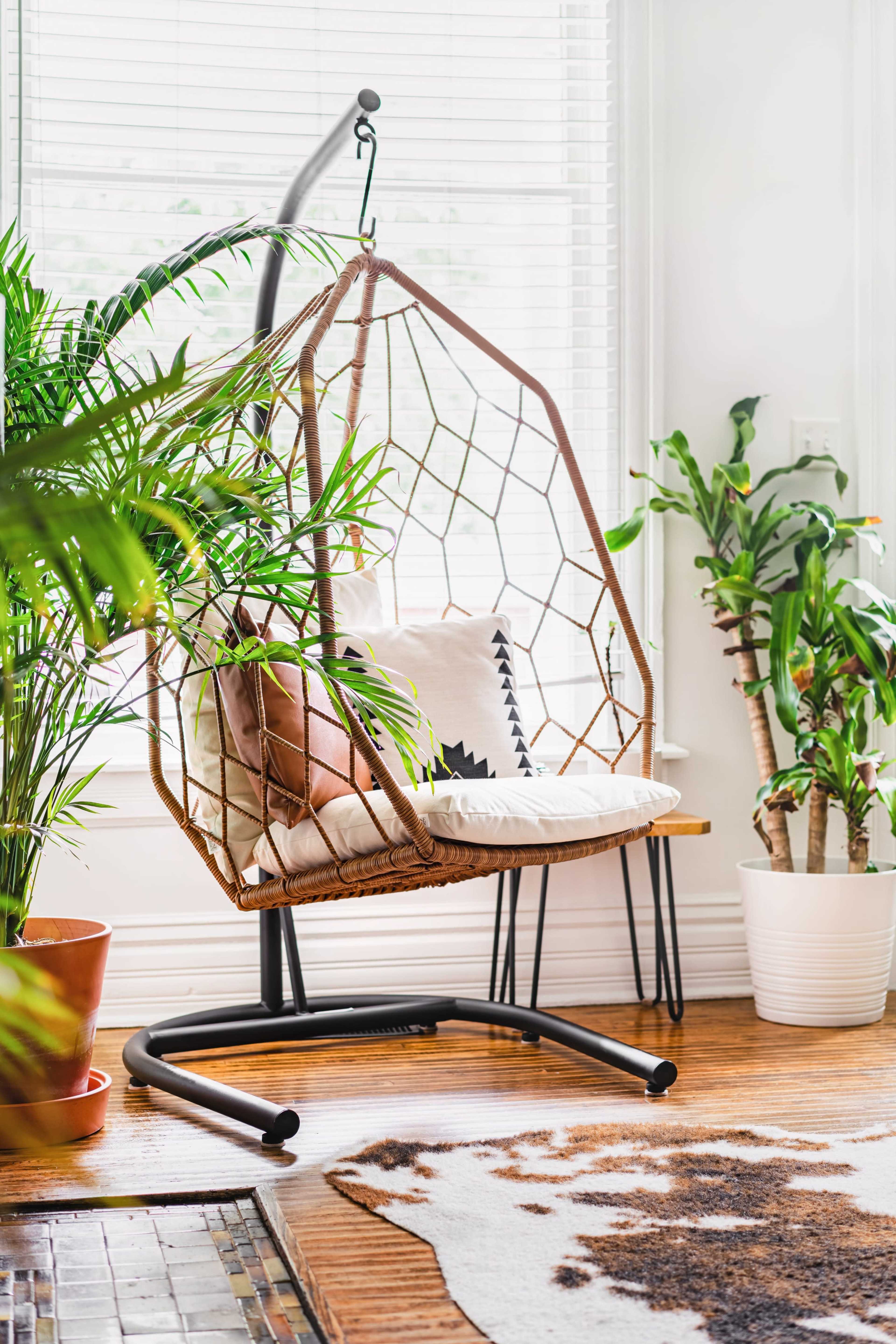 A hanging chair with a cushion is positioned near large potted plants in a bright, airy room.