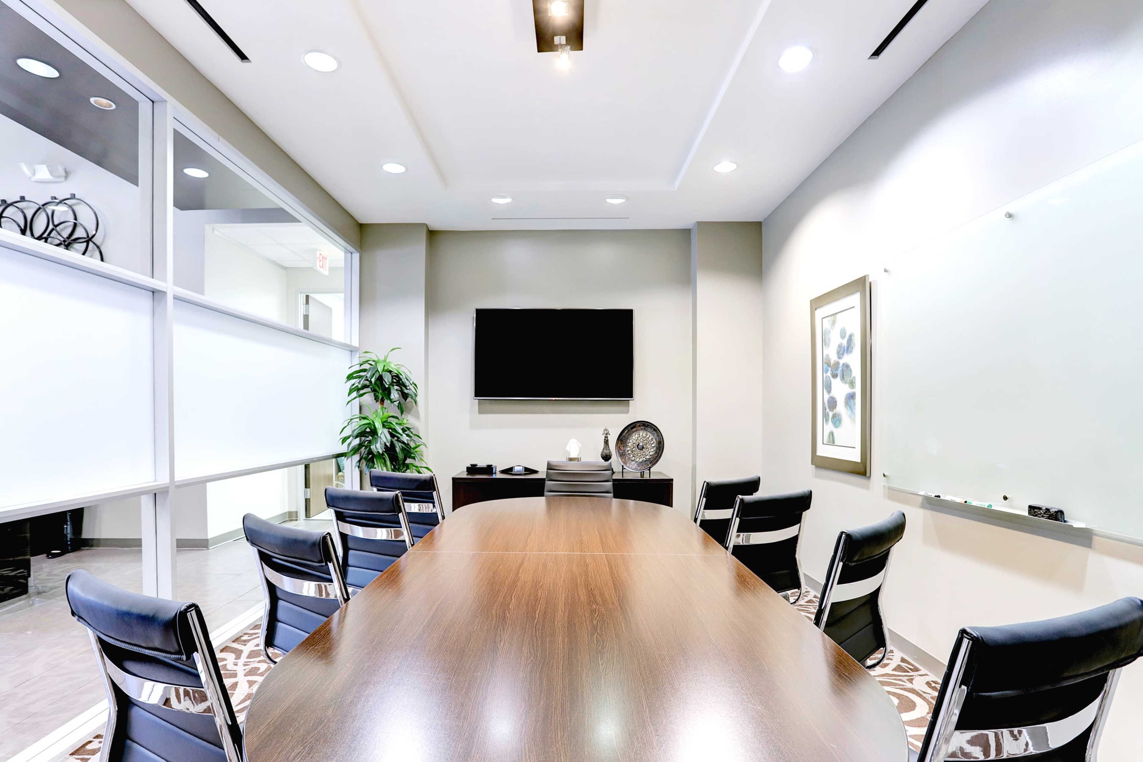 A modern conference room features a large wooden table surrounded by black chairs, with a television, whiteboard, and decorative elements on the walls.