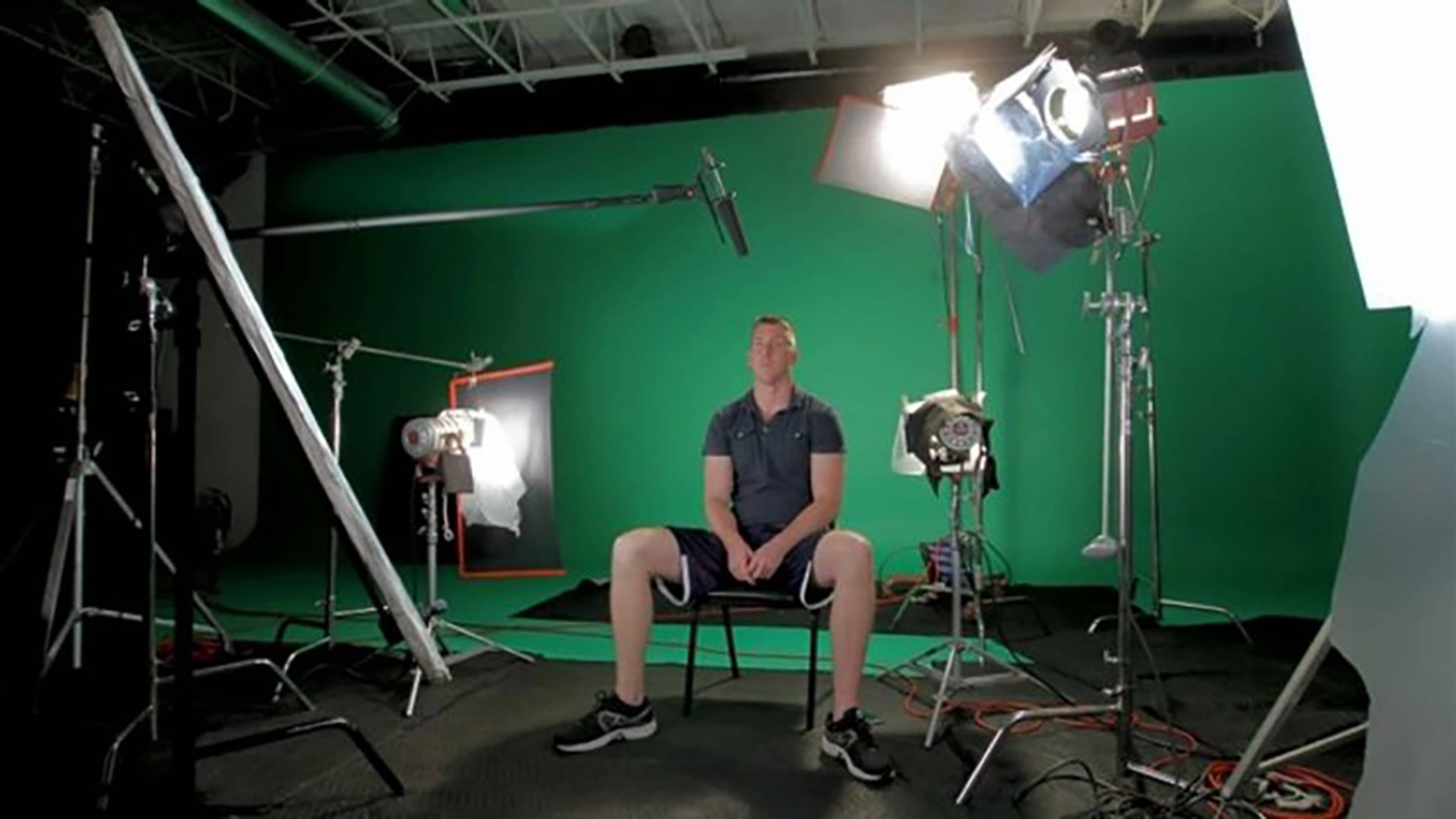 A man sits on a chair in front of a green screen setup, surrounded by studio lights and equipment.