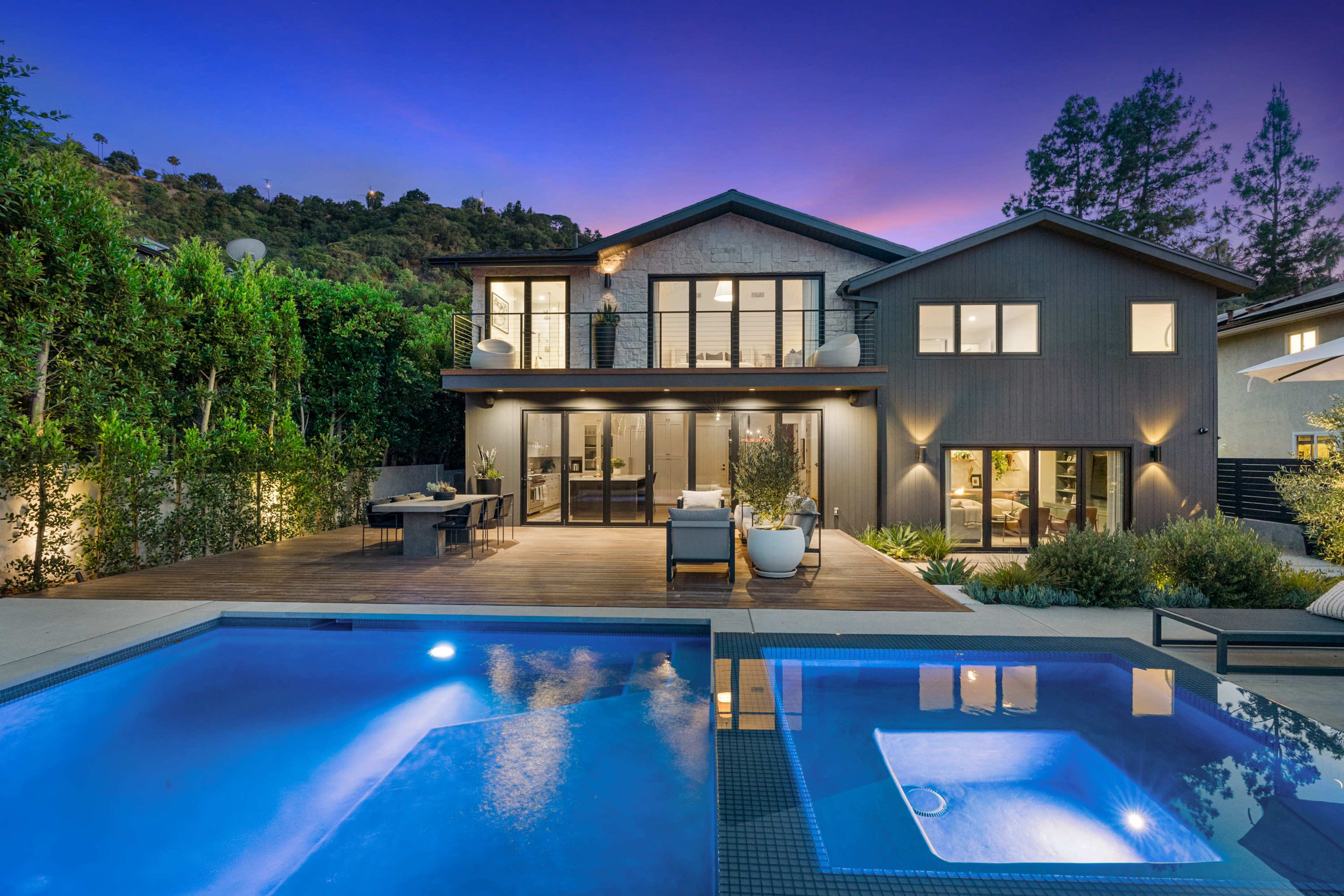 A modern two-story house with large glass doors overlooks a pool area surrounded by lush greenery and outdoor seating at twilight.