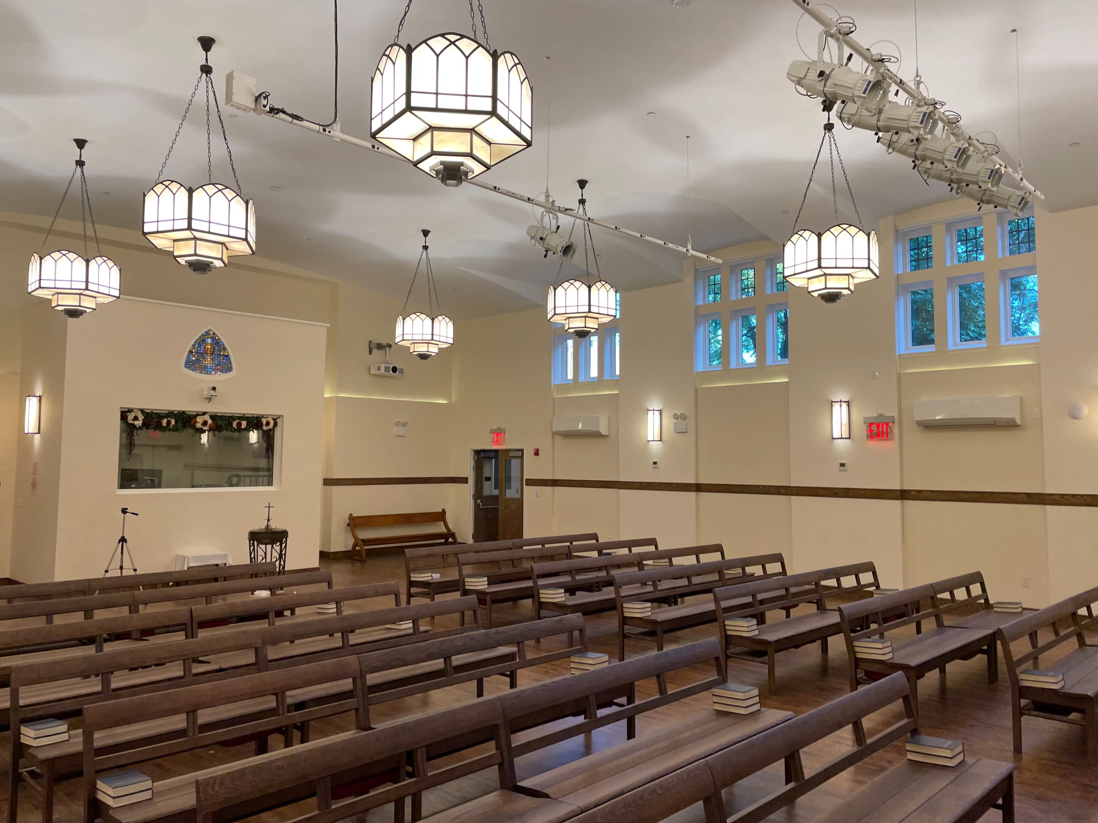 An interior of a spacious, well-lit hall with rows of wooden benches, decorative pendant lights, and large windows.