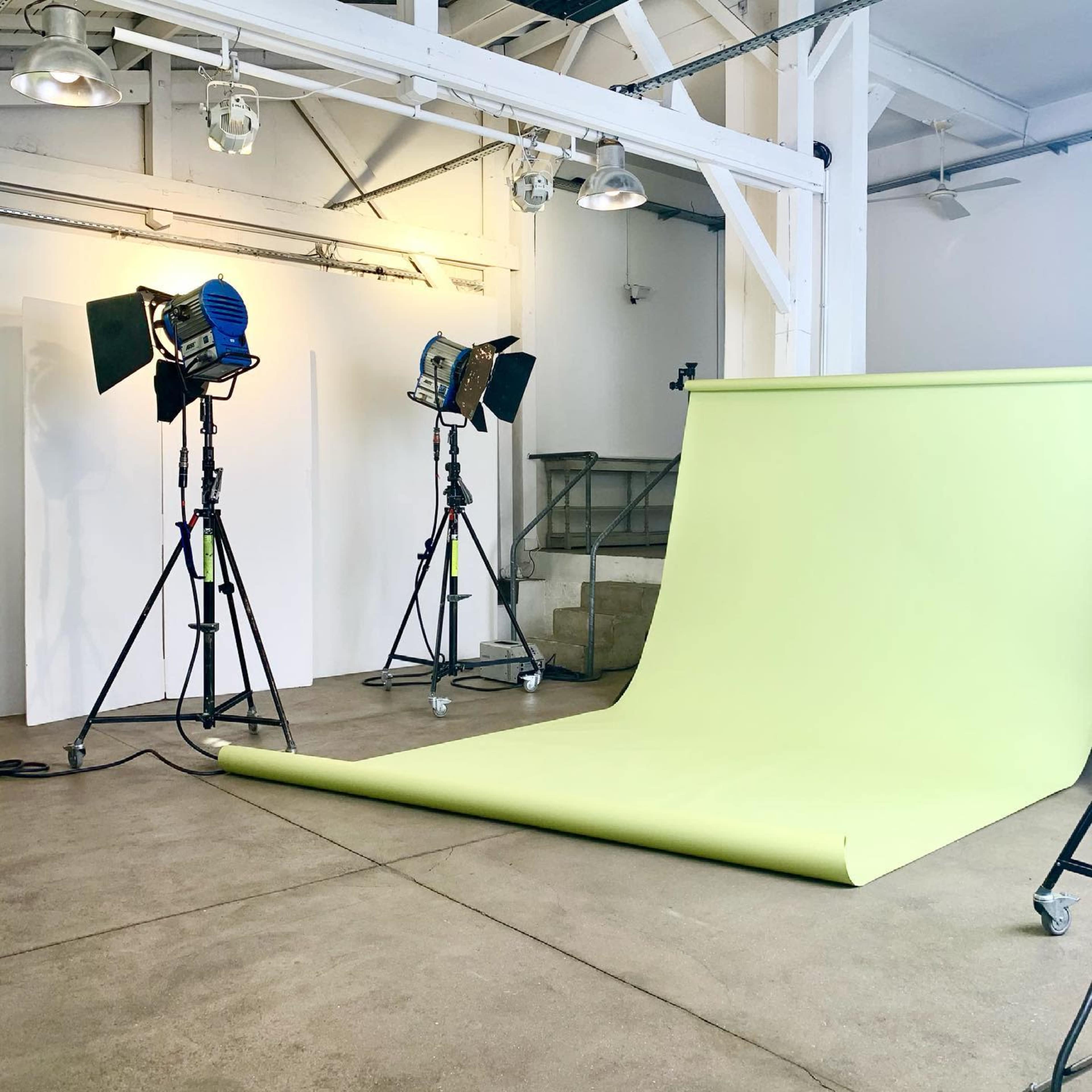 A photo studio with a light green backdrop, three lighting setups, and a partially exposed staircase in the background.