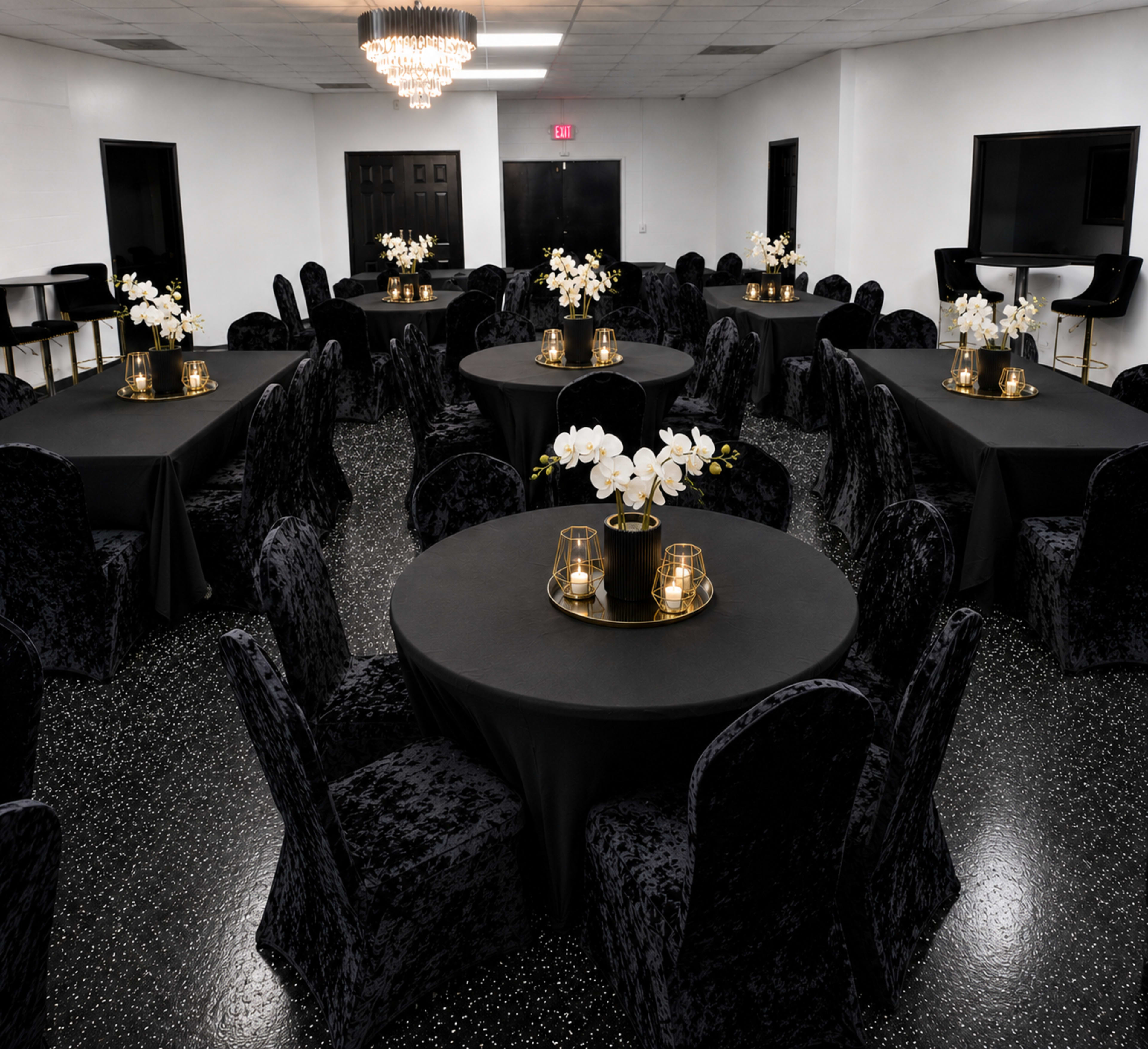 The image shows a banquet hall set up with several round tables covered in black tablecloths, each adorned with floral centerpieces and candles, surrounded by plush black chairs.