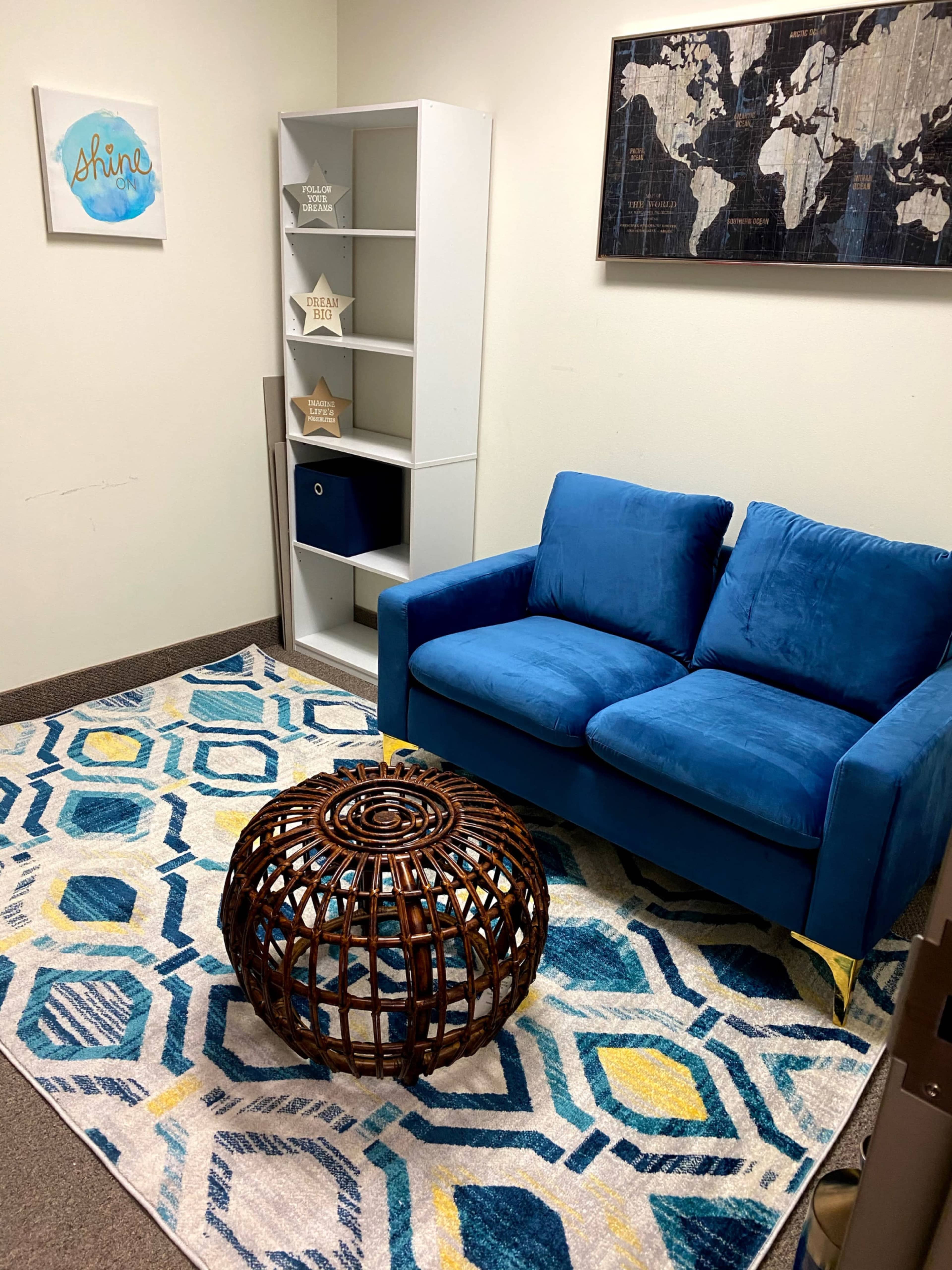 A blue couch sits on a patterned rug next to a white bookshelf and a round wicker coffee table in a small, simple room.
