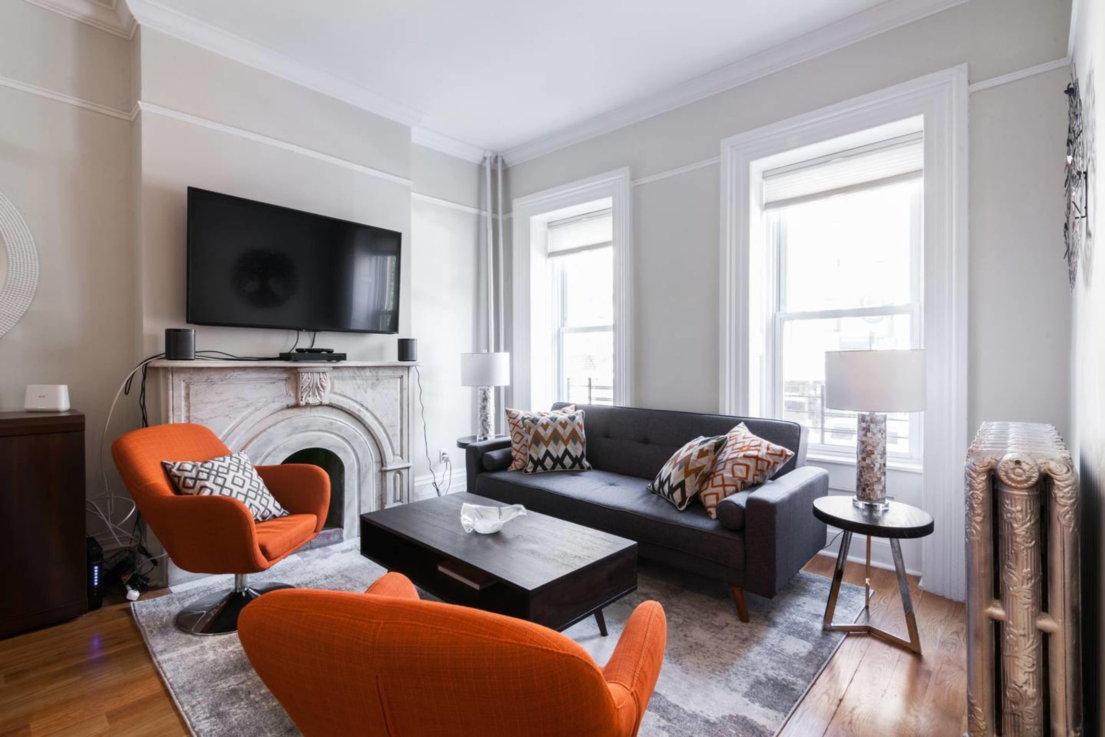 The living room features a gray couch, two orange armchairs, a dark wood coffee table, and a marble fireplace, with a television mounted on the wall above it.