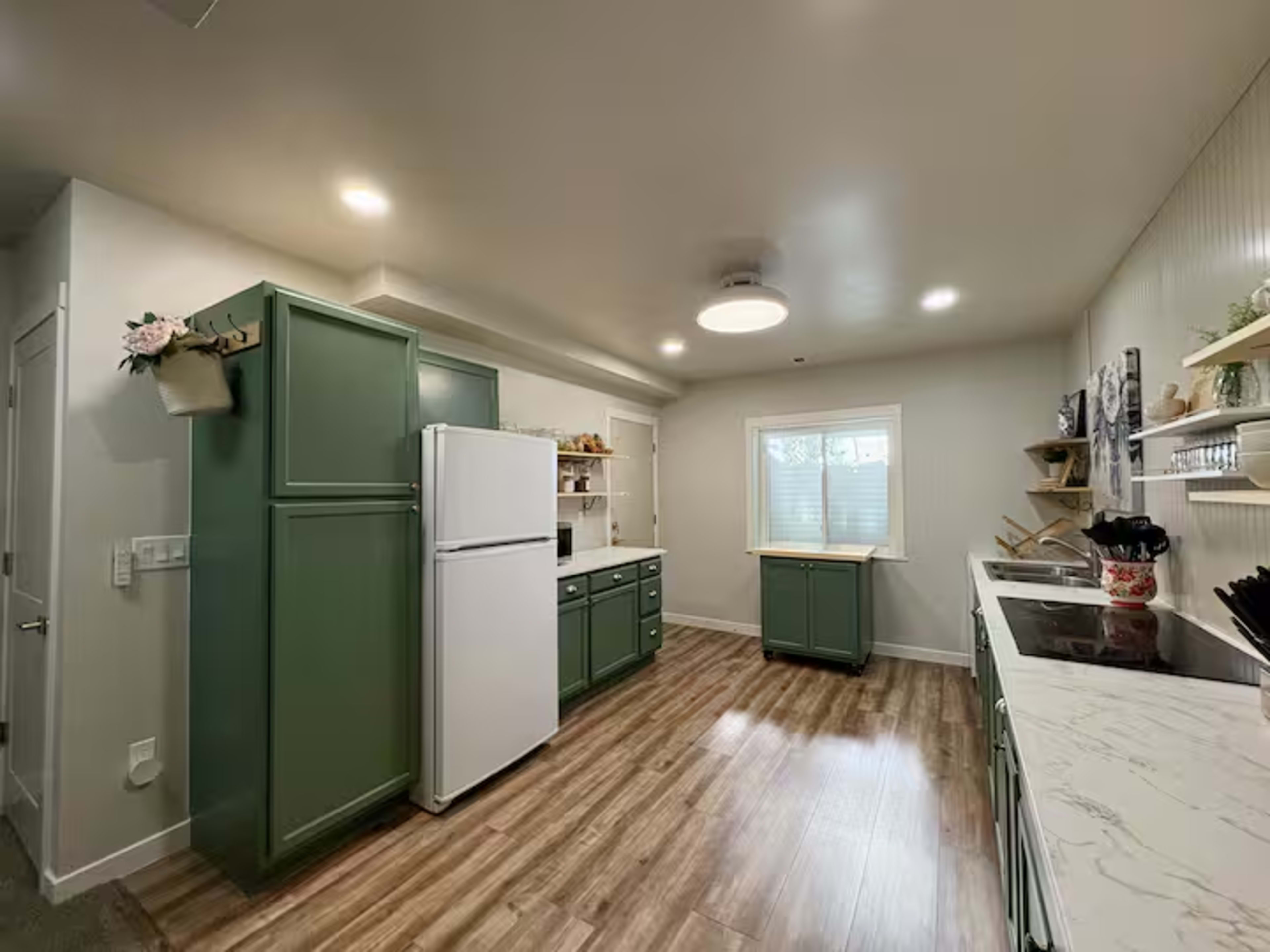The image shows a modern kitchen with green cabinetry, a white refrigerator, and light-colored countertops, featuring a window that lets in natural light.