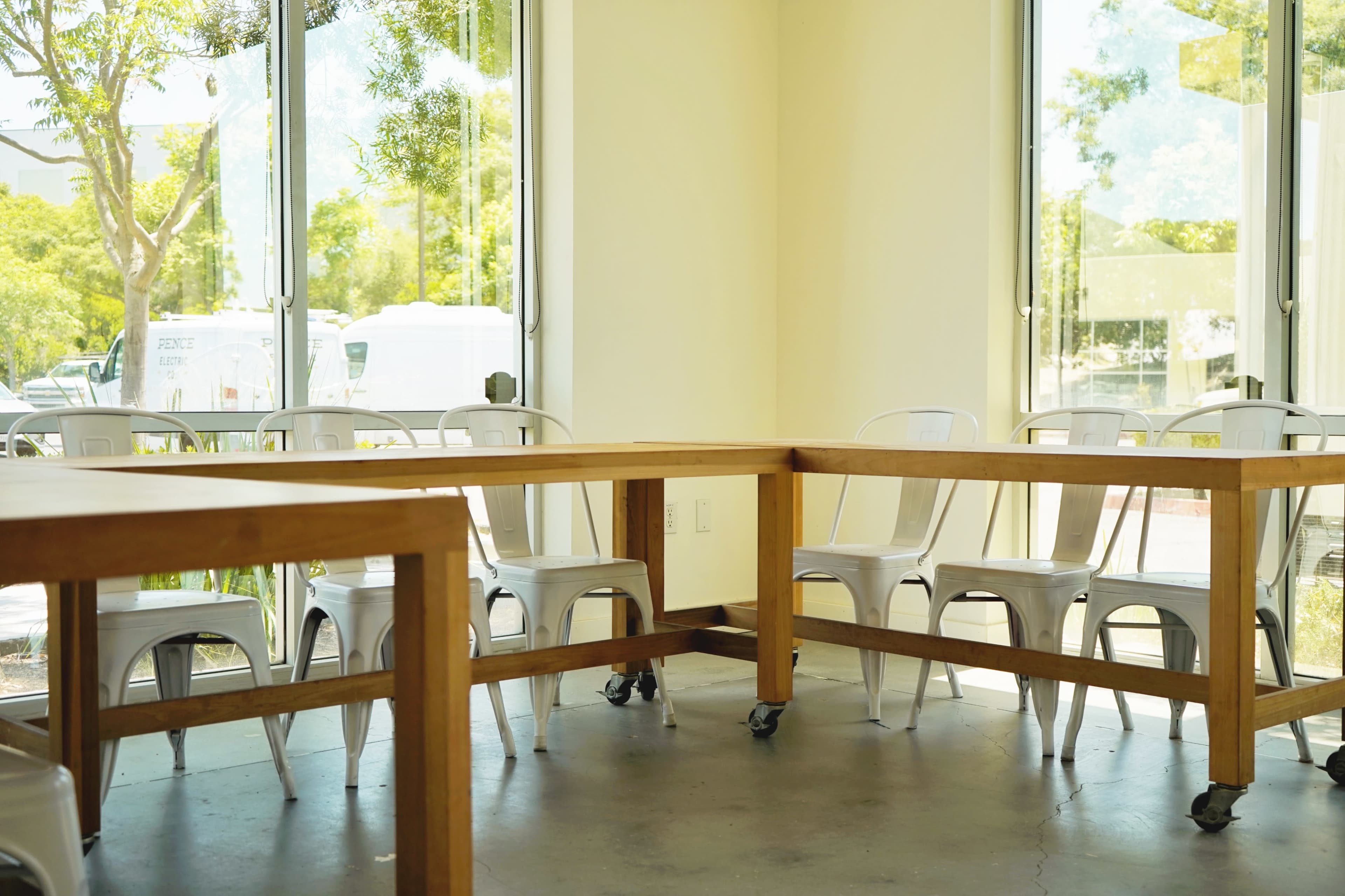A spacious room features large wooden tables surrounded by metal chairs, with large windows letting in natural light.