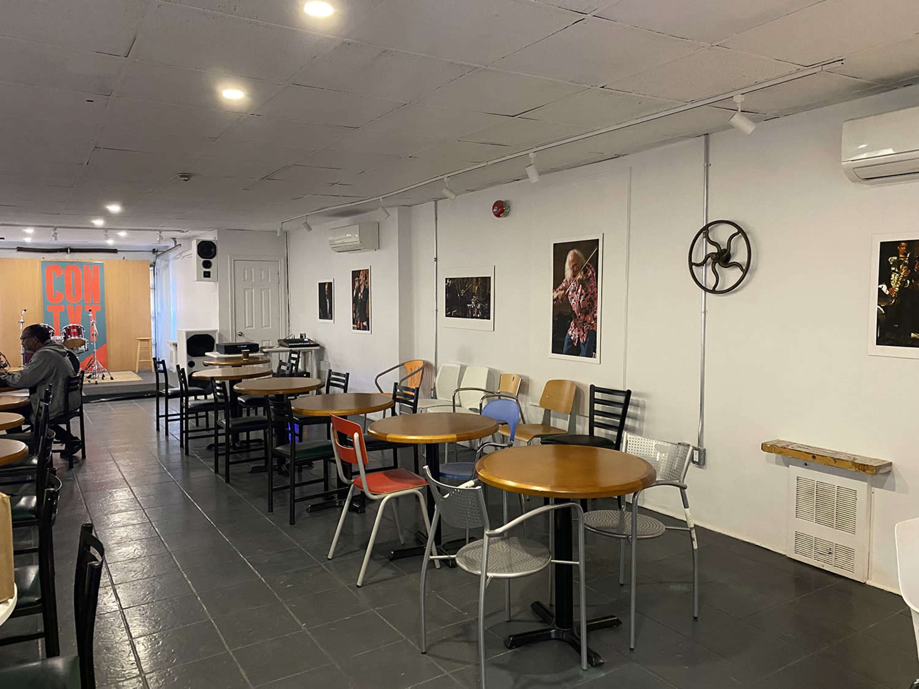 The image shows a spacious café with several tables and chairs arranged around the room, featuring wall art and a small stage at one end.