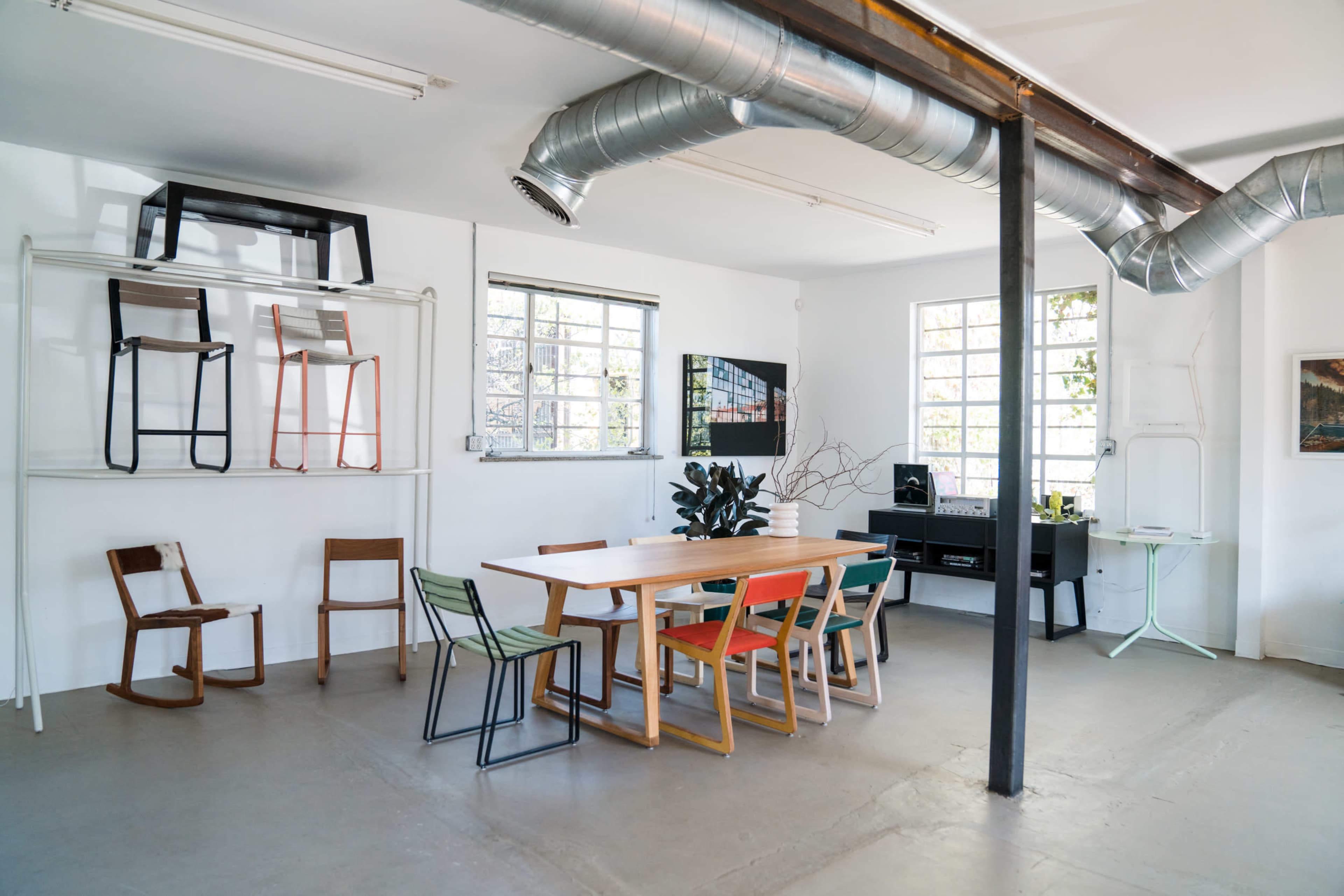 A modern, minimalist design studio features a large wooden table surrounded by colorful chairs, with additional chairs displayed on shelves and large windows allowing natural light.