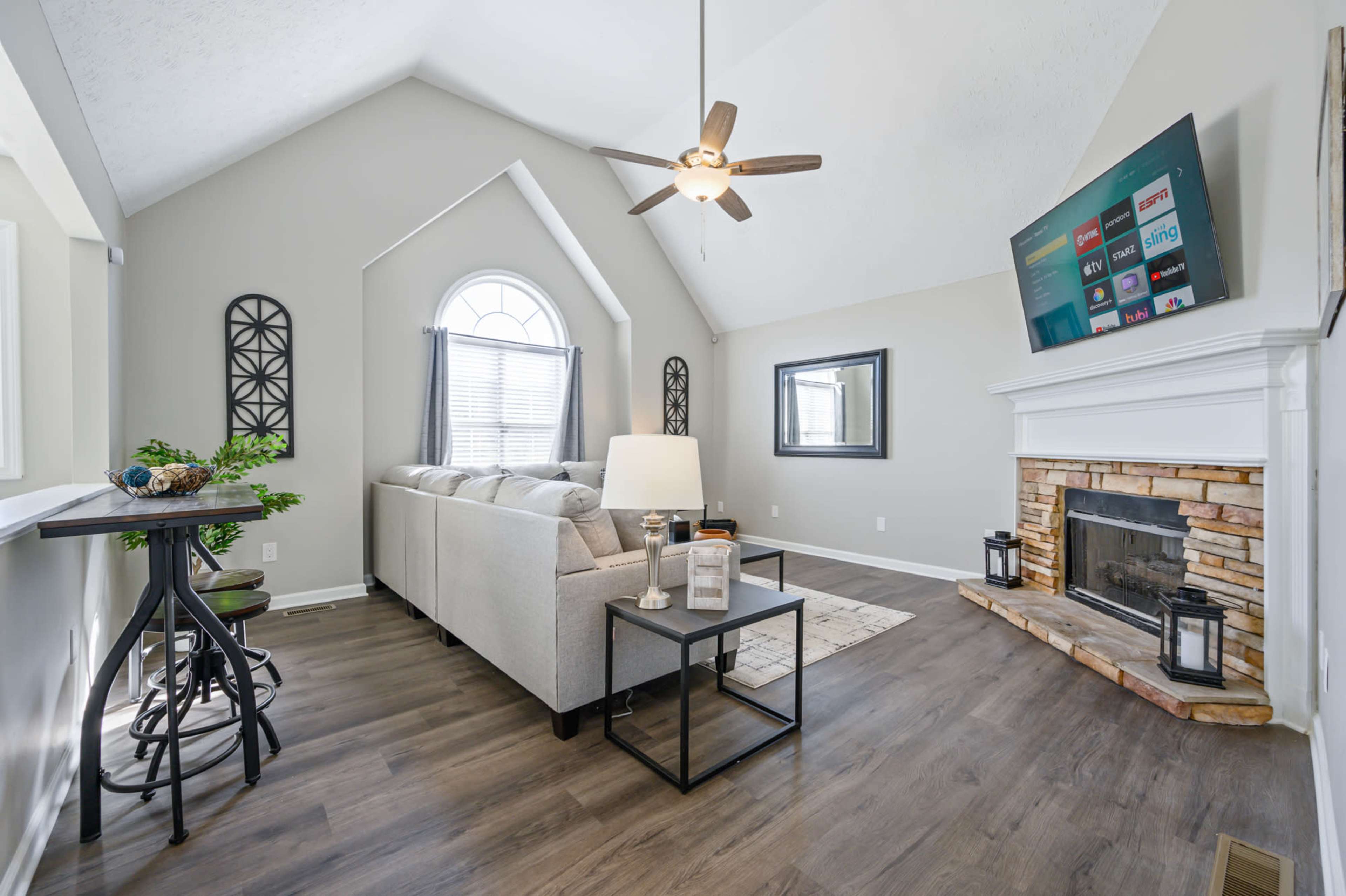 A modern living room features a light gray sofa, a coffee table, and a stone fireplace beneath a wall-mounted television, with large windows allowing natural light.