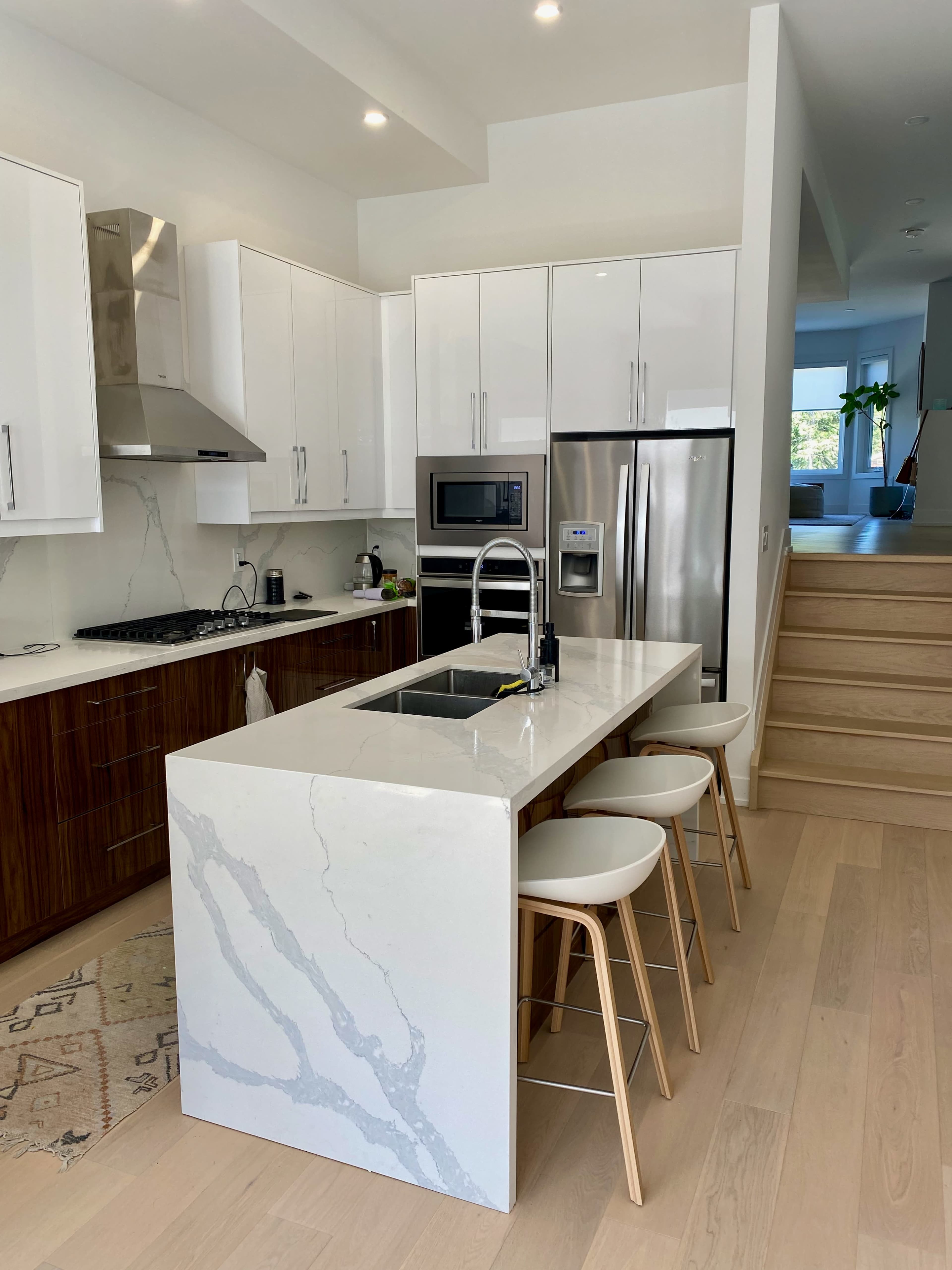 A modern kitchen features a central island with a sink, surrounded by three stools, and stainless steel appliances against white cabinetry.