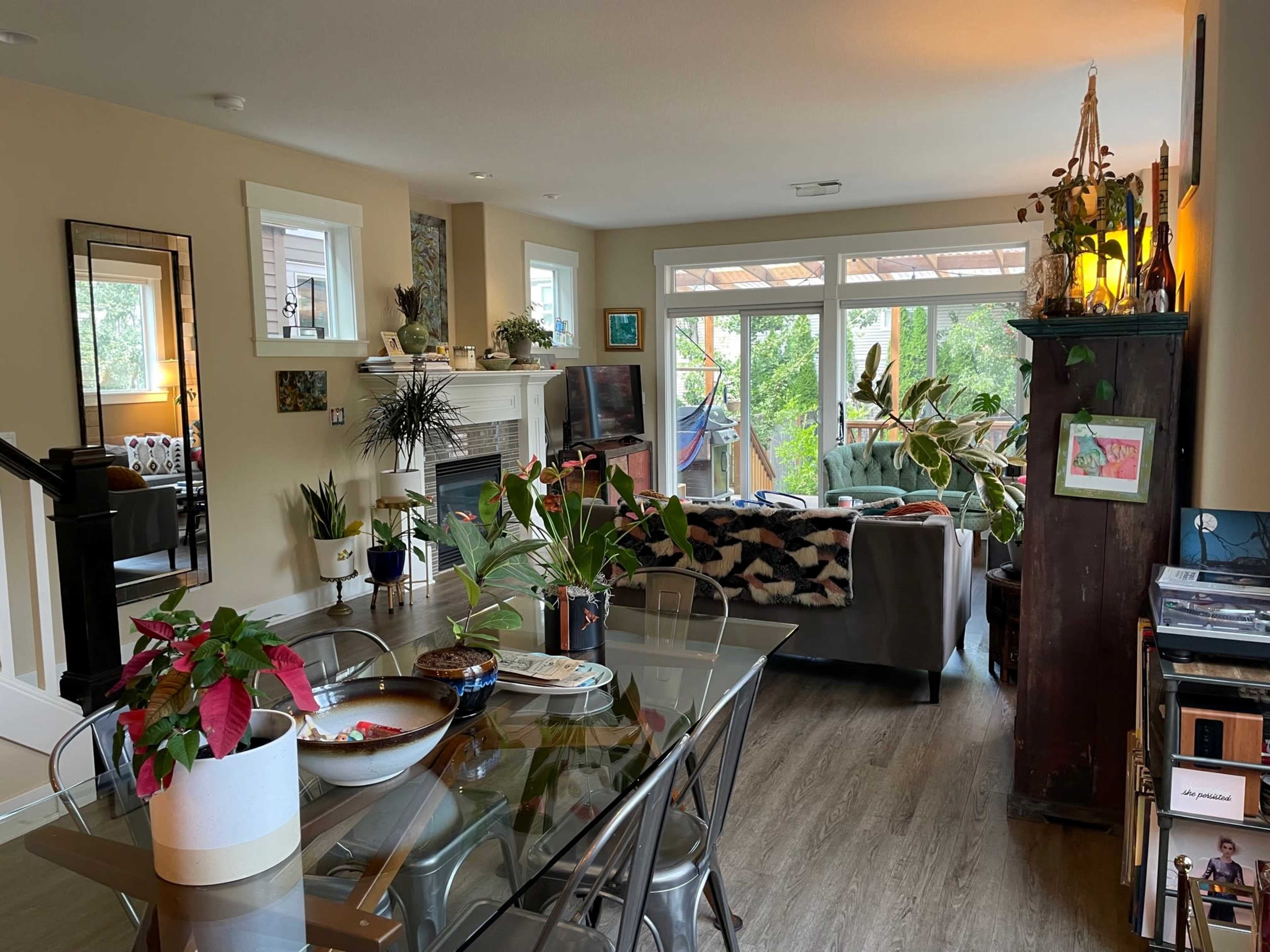 A brightly-lit living room features a dining table, a cozy seating area, plants, and large windows with a view of greenery outside.