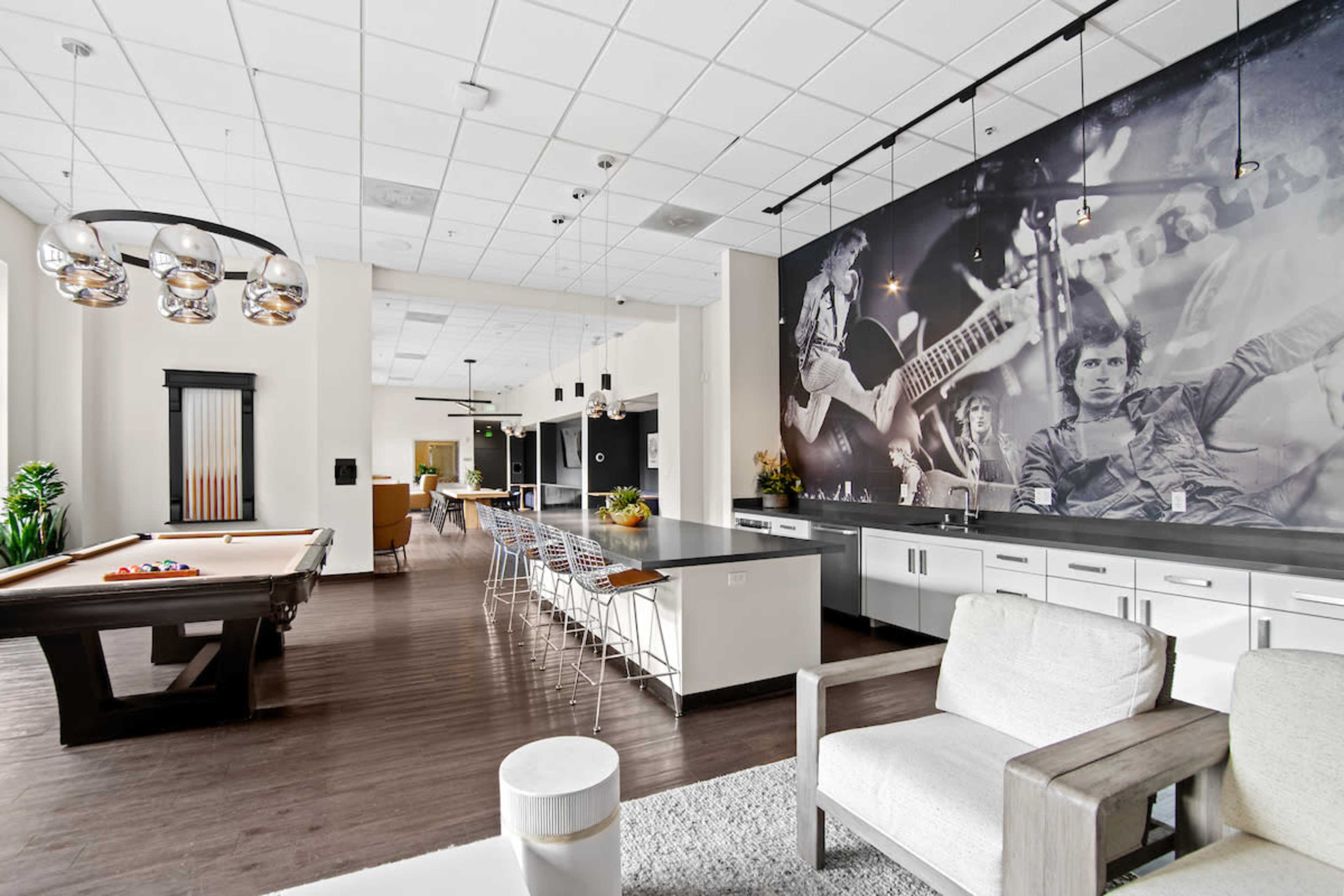 The image shows a modern recreational room with a pool table, a kitchen island with bar stools, and a large mural on the wall featuring musicians.