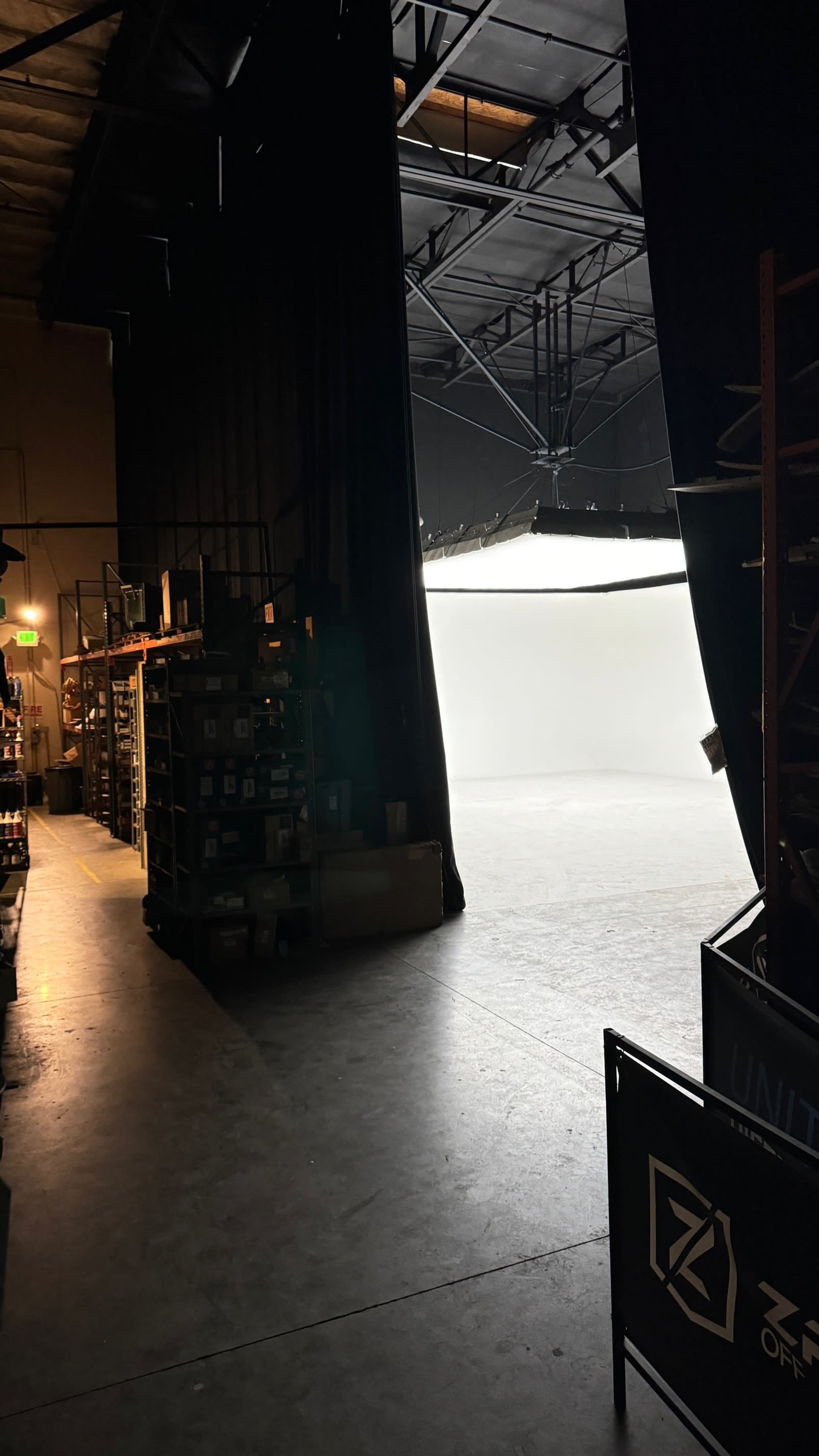 The image shows a dimly lit warehouse area with shelves on one side and a brightly lit blank backdrop visible at the far end through an open curtain.