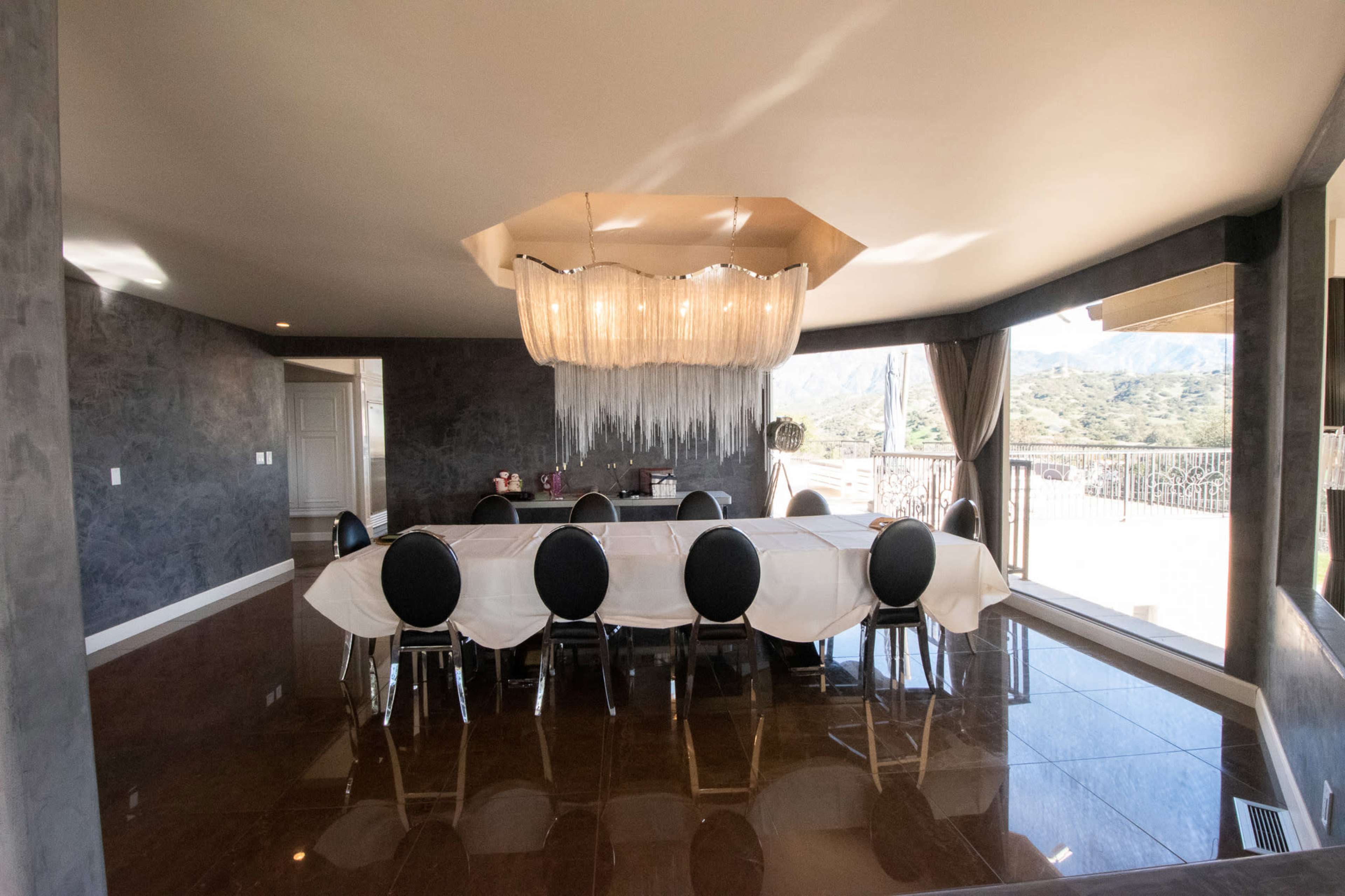 A spacious dining room features a large table surrounded by black chairs under a unique chandelier, with large windows revealing a mountainous backdrop.