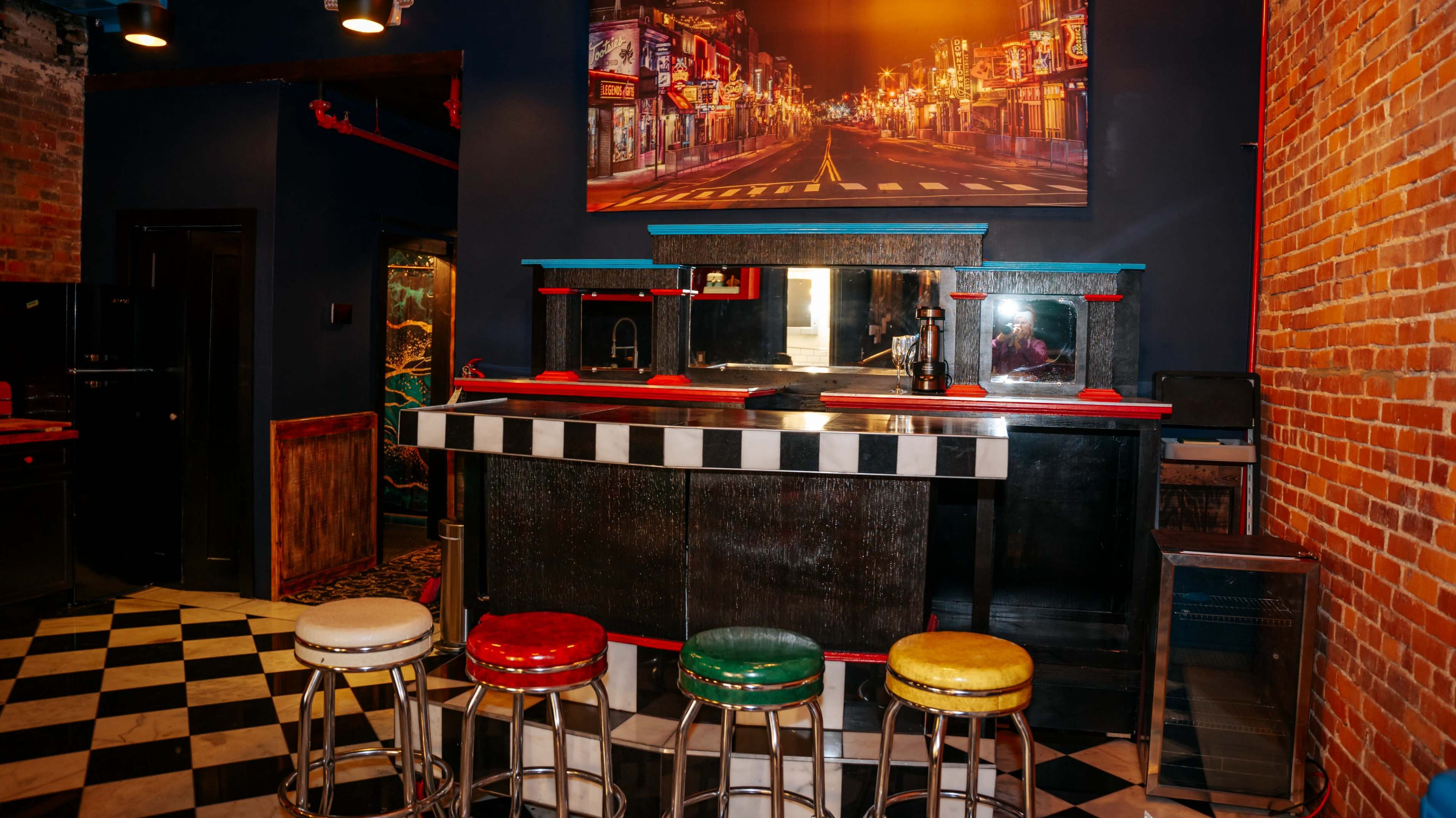 The image shows a bar area with a checkerboard floor, colorful stools, and a large wall-mounted photo of a street scene.