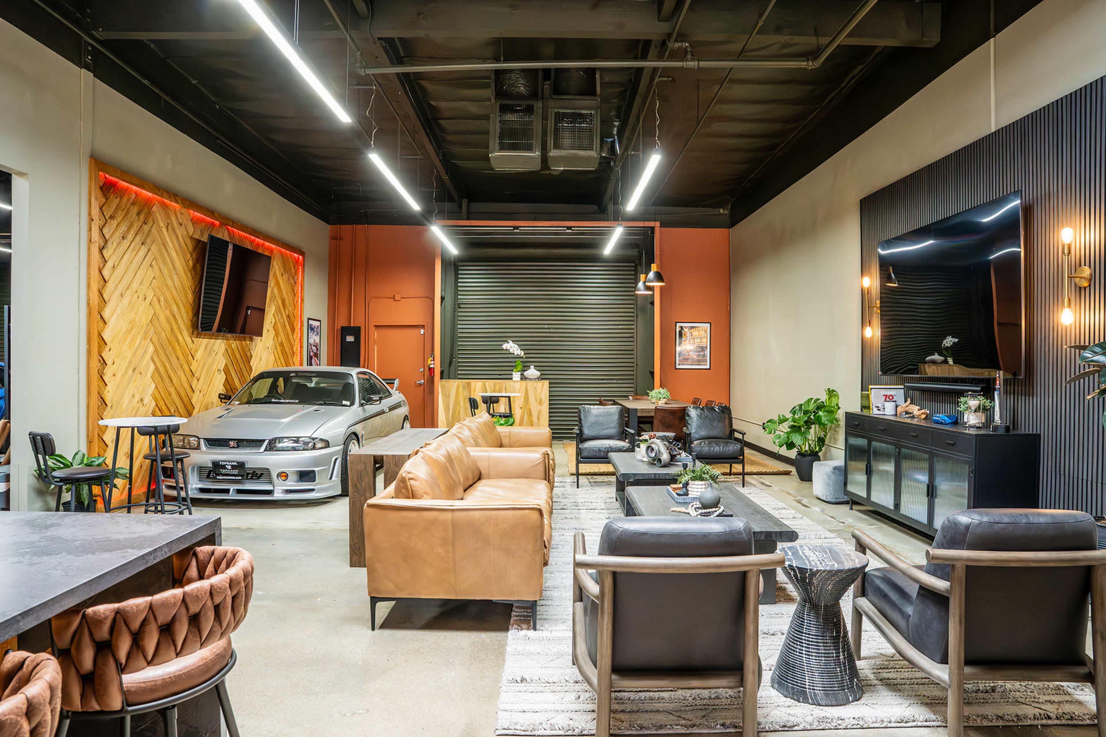 The image shows a modern indoor space that combines a lounge area with leather sofas, a coffee table, and a parked sports car, featuring decorative plants and wall-mounted TVs.