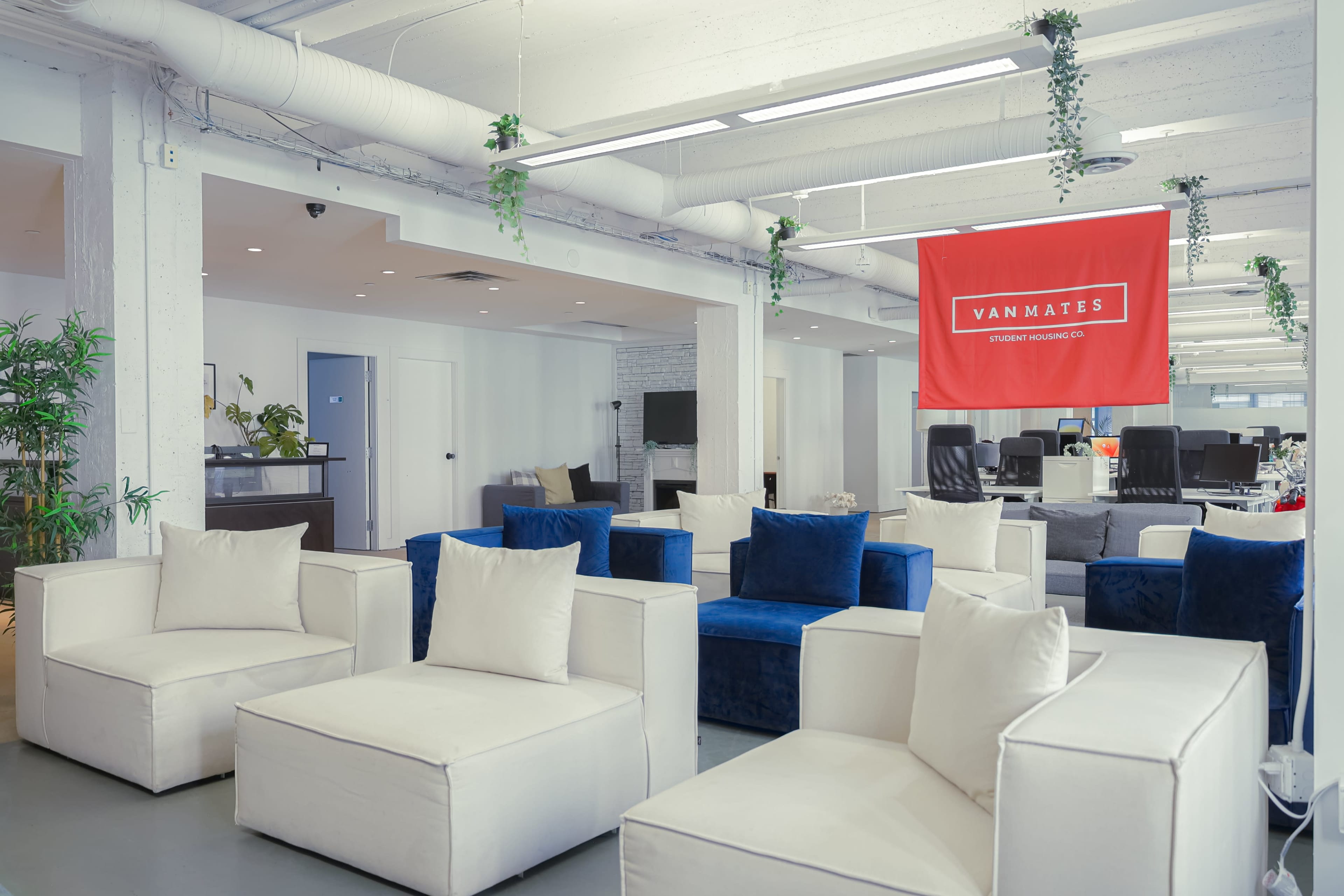 The image shows a modern coworking space with white and blue seating arranged around a central area, featuring a red banner that reads "VANMATES."