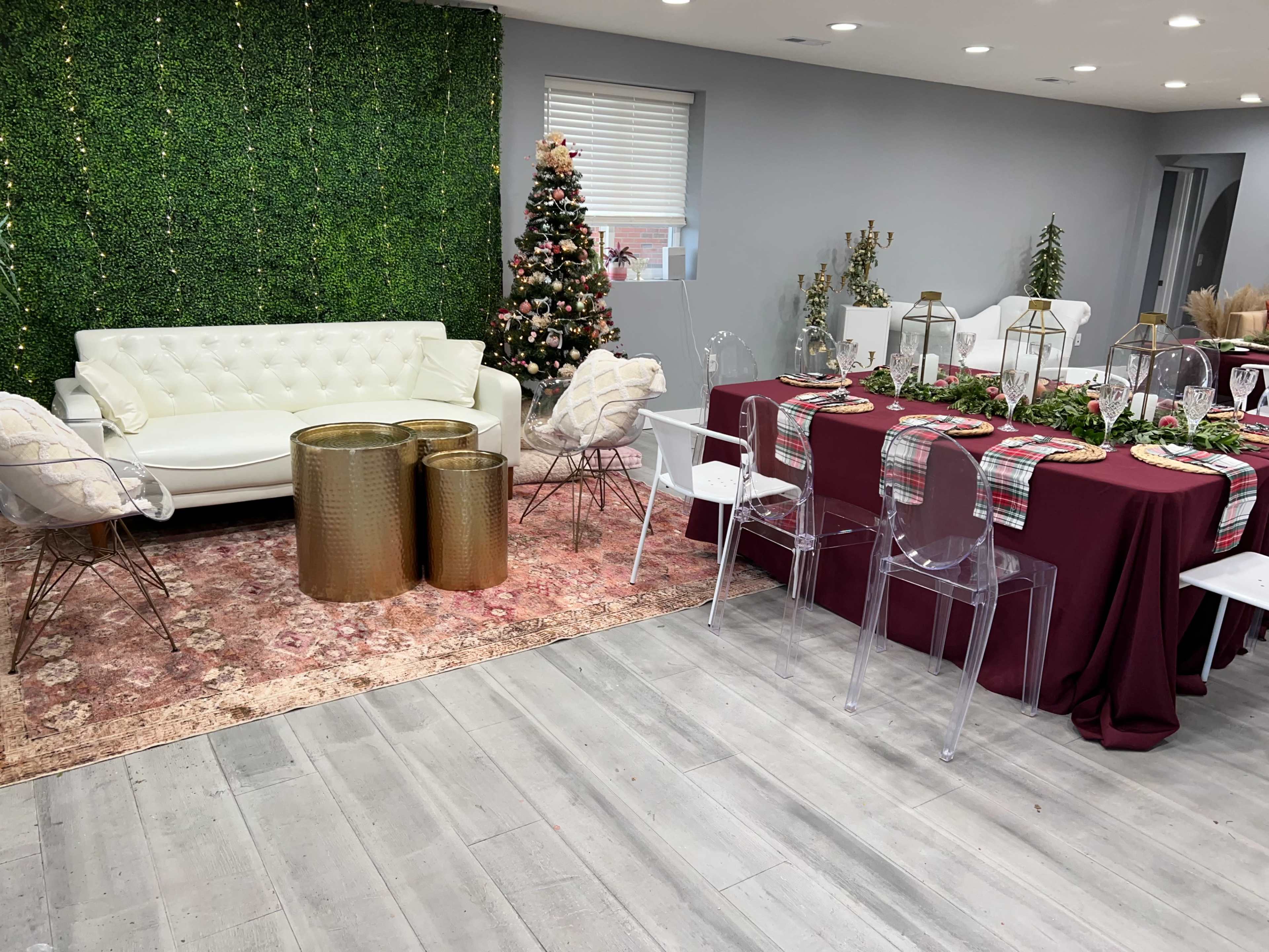 A decorated event space featuring a burgundy dining table set, transparent chairs, a white couch, and a Christmas tree in the background.