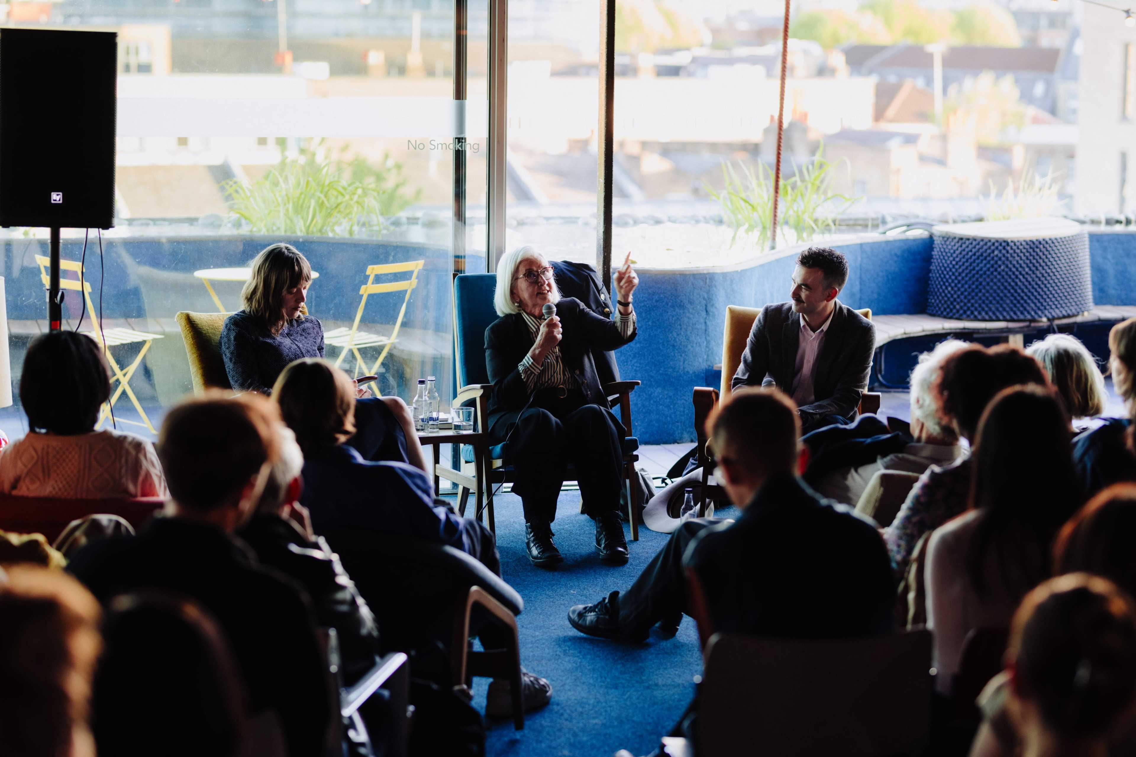 A panel discussion takes place in a well-lit room with large windows, featuring three speakers seated in chairs while an audience attentively listens.