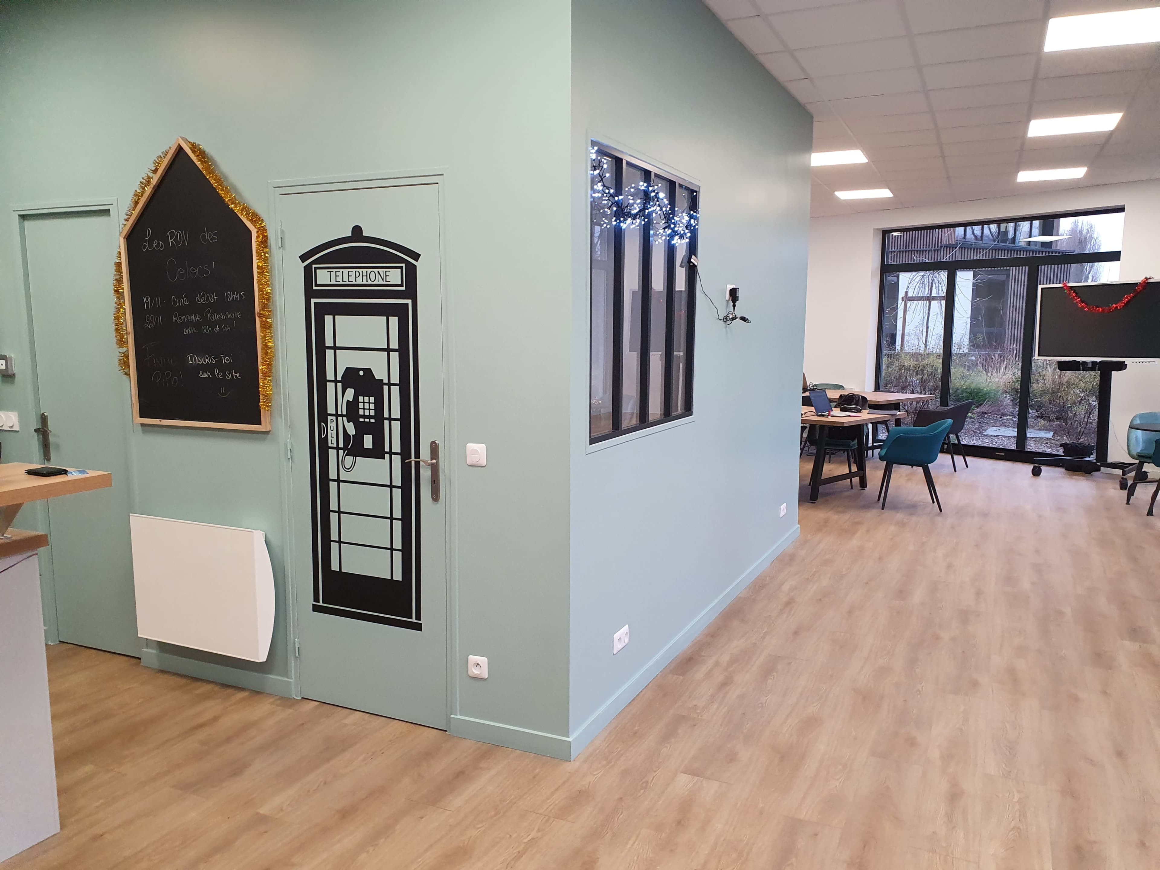 A modern office space with a green wall featuring a decorative telephone booth design, a chalkboard, and a view of tables and chairs in the background.