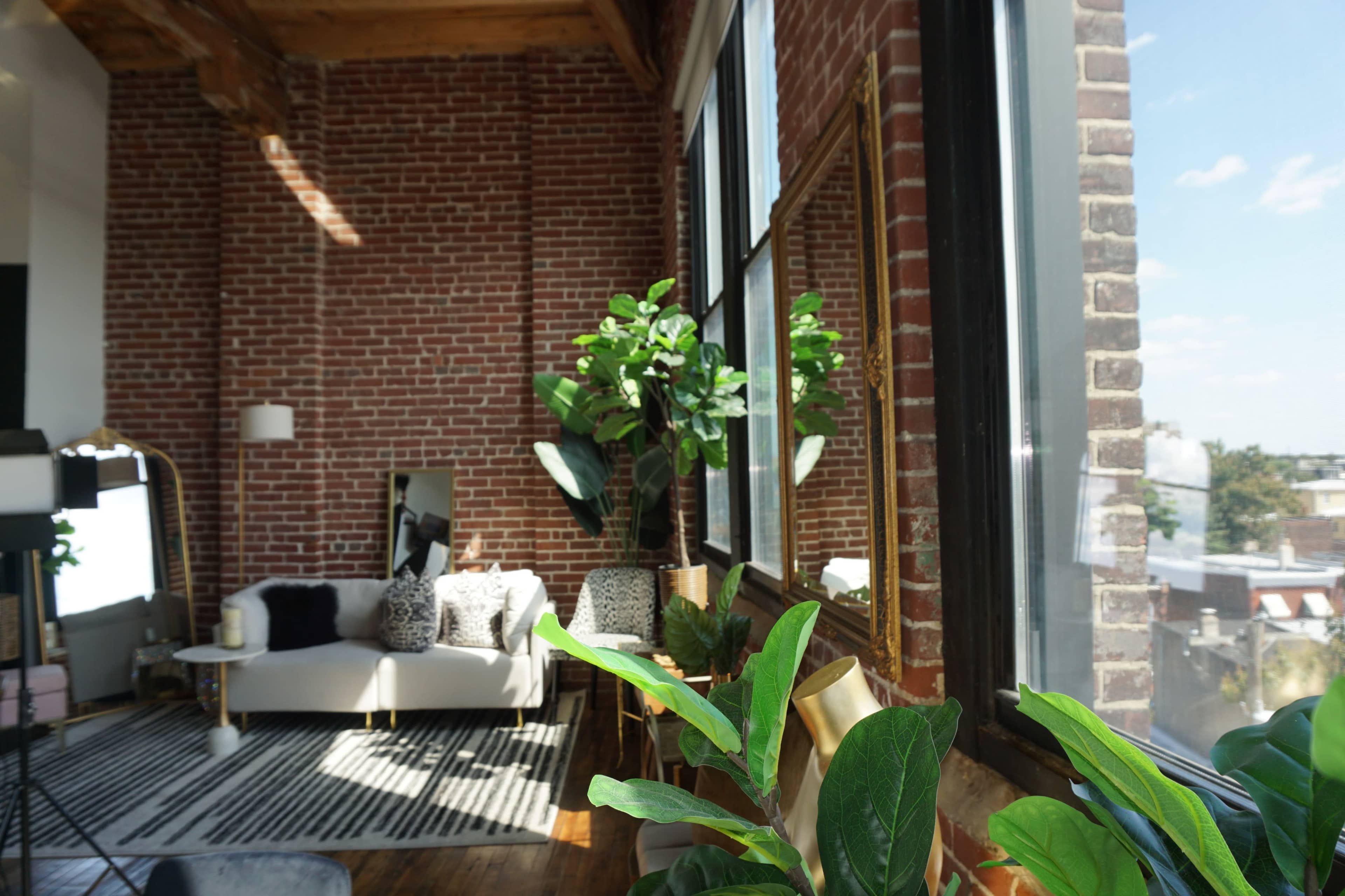 A bright living area features a white sofa with patterned pillows, large windows, and a brick wall backdrop adorned with indoor plants.