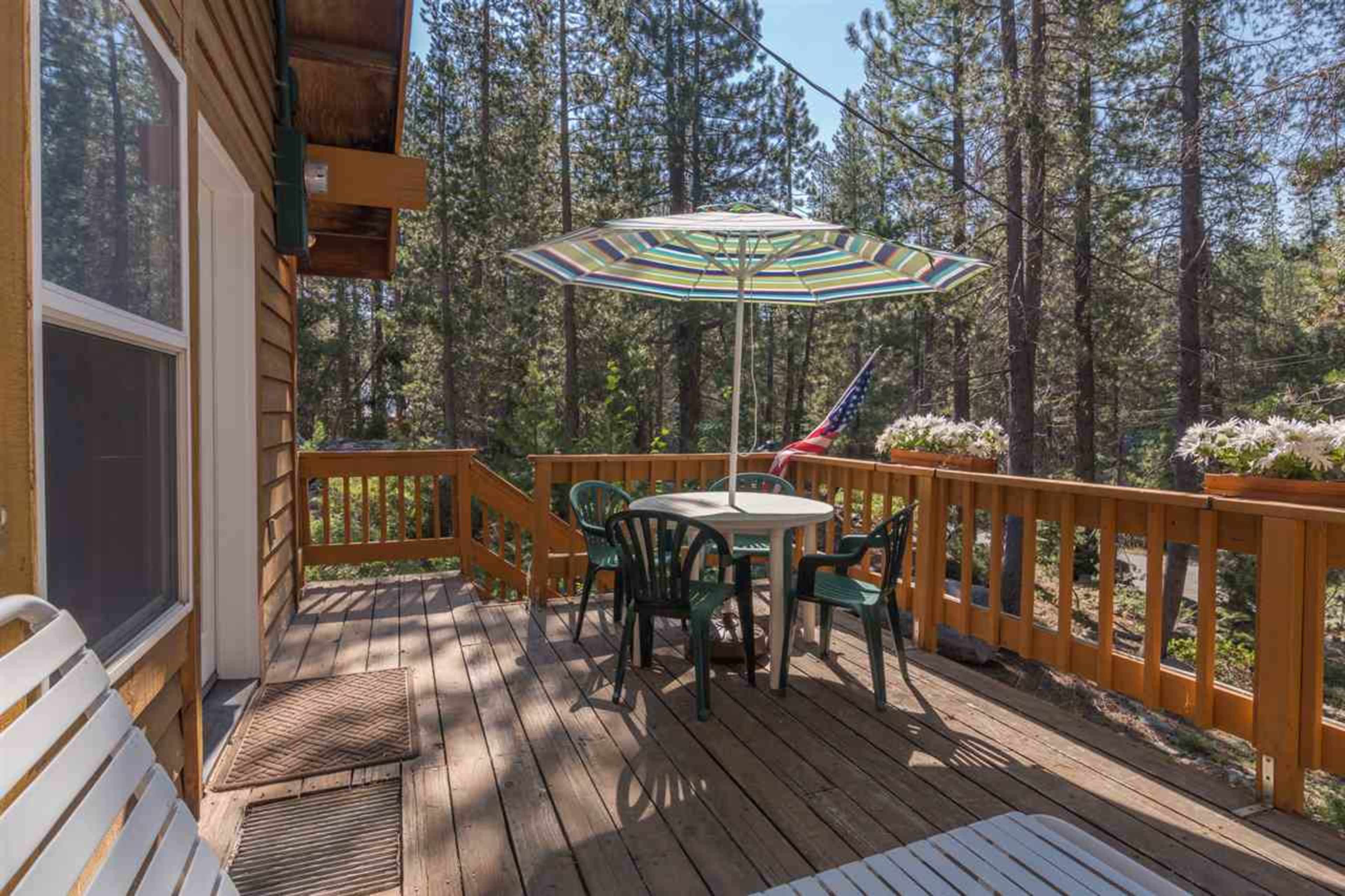 A wooden deck features a round table with chairs and a striped umbrella, surrounded by tall trees and shrubs.
