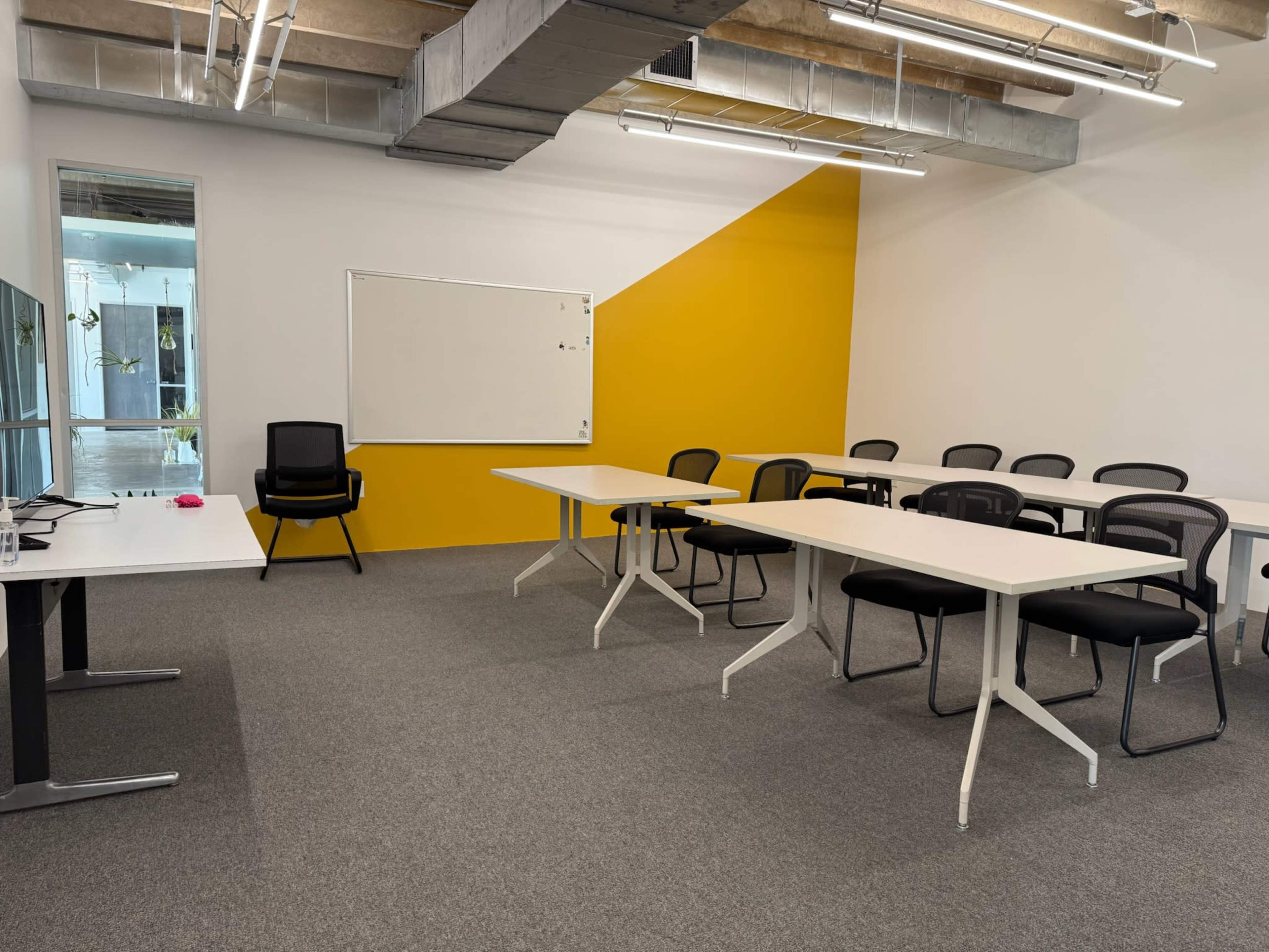 The photo shows a modern meeting room with multiple tables arranged in a U-shape, black chairs, a whiteboard, and a yellow accent wall.