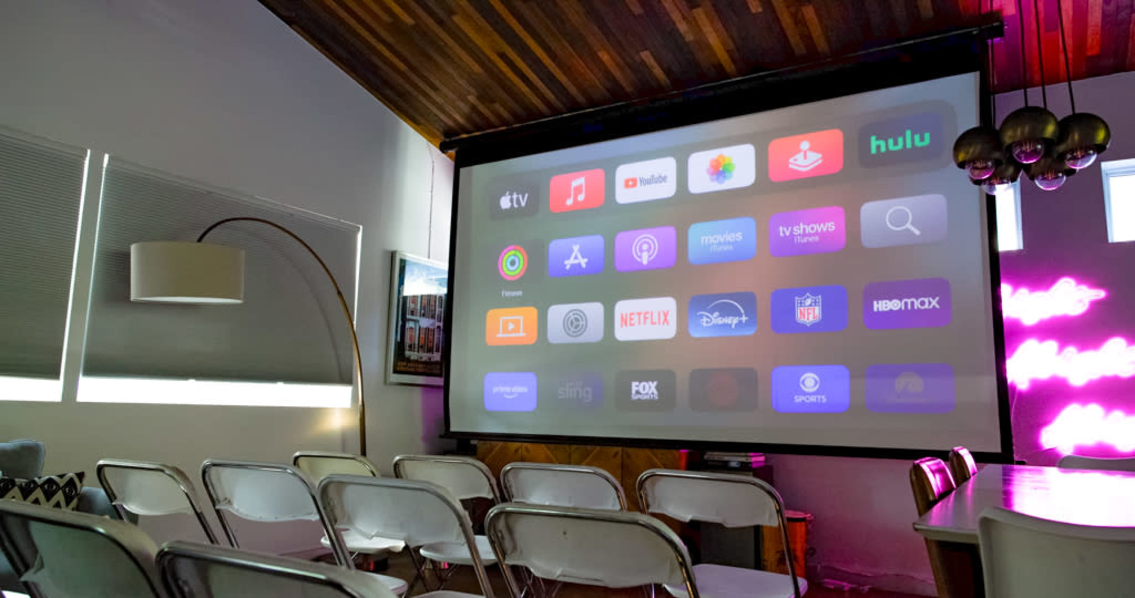 The image shows a home theater setup with a large projection screen displaying various streaming service icons, surrounded by rows of white folding chairs.
